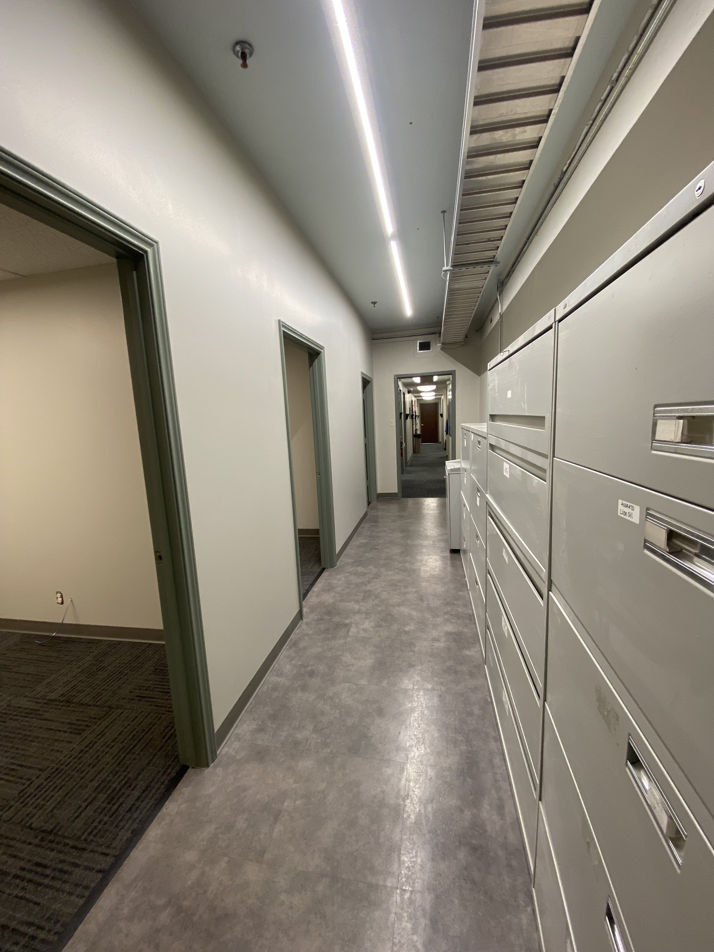 Empty office hallway with multiple doors, gray flooring, and wall-mounted mailboxes on the right side.