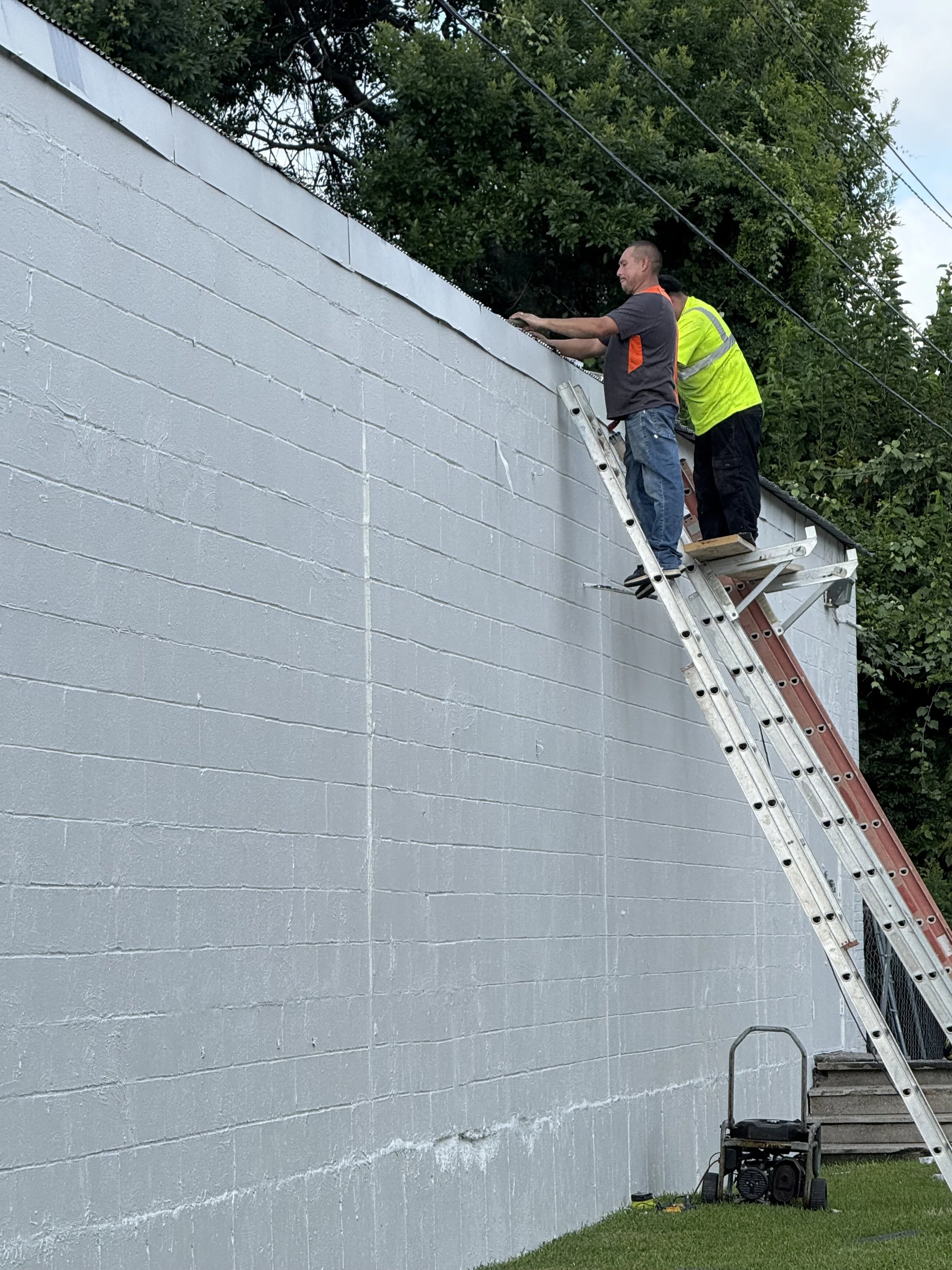 Two men are on ladders working on the top edge of a white cinder block wall, with one man wearing a gray shirt and the other in a yellow safety vest. There are green trees and power lines in the background.