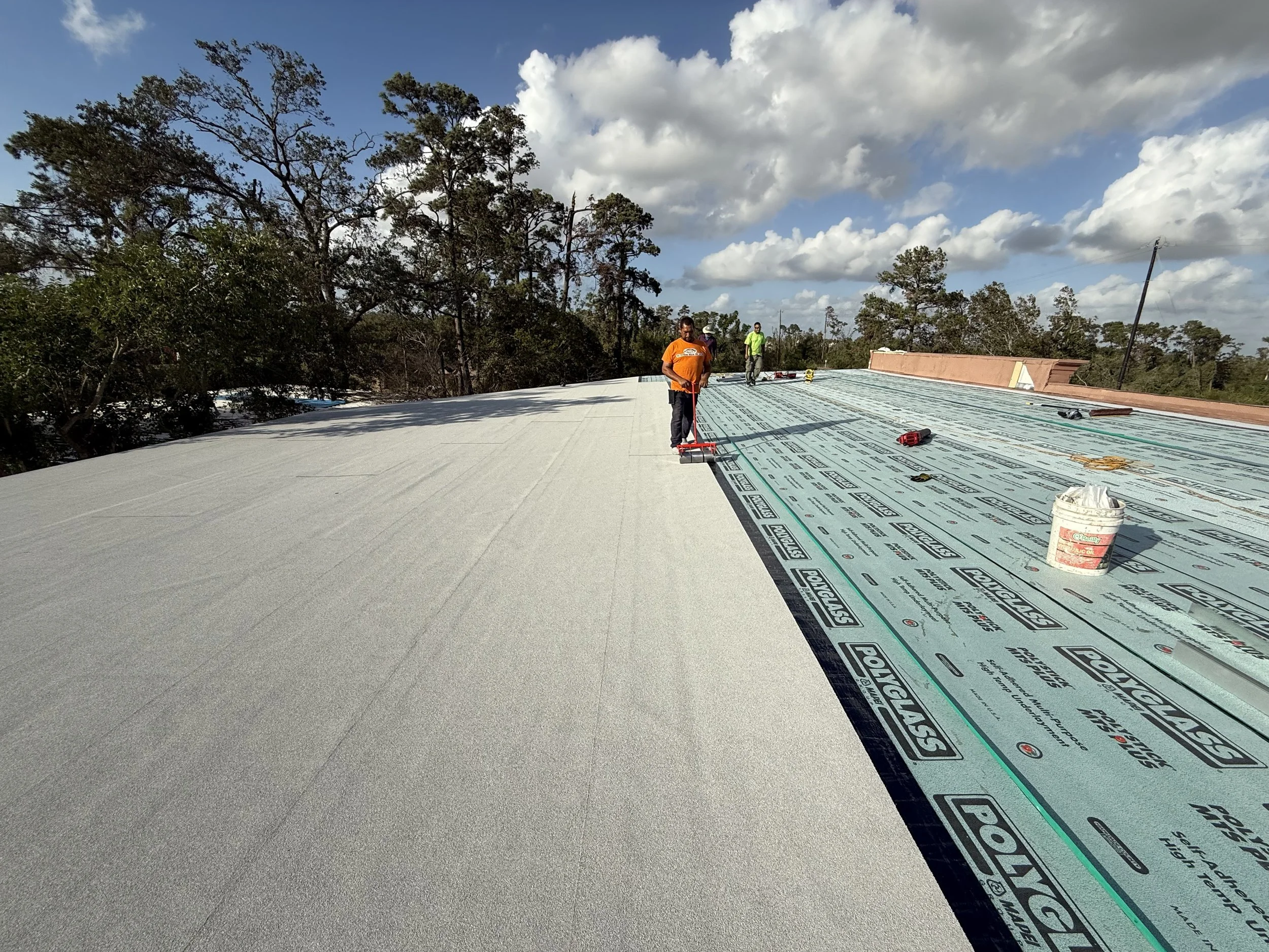 Construction workers installing a roof with a weatherproof underlayment, under a partly cloudy sky, surrounded by trees.