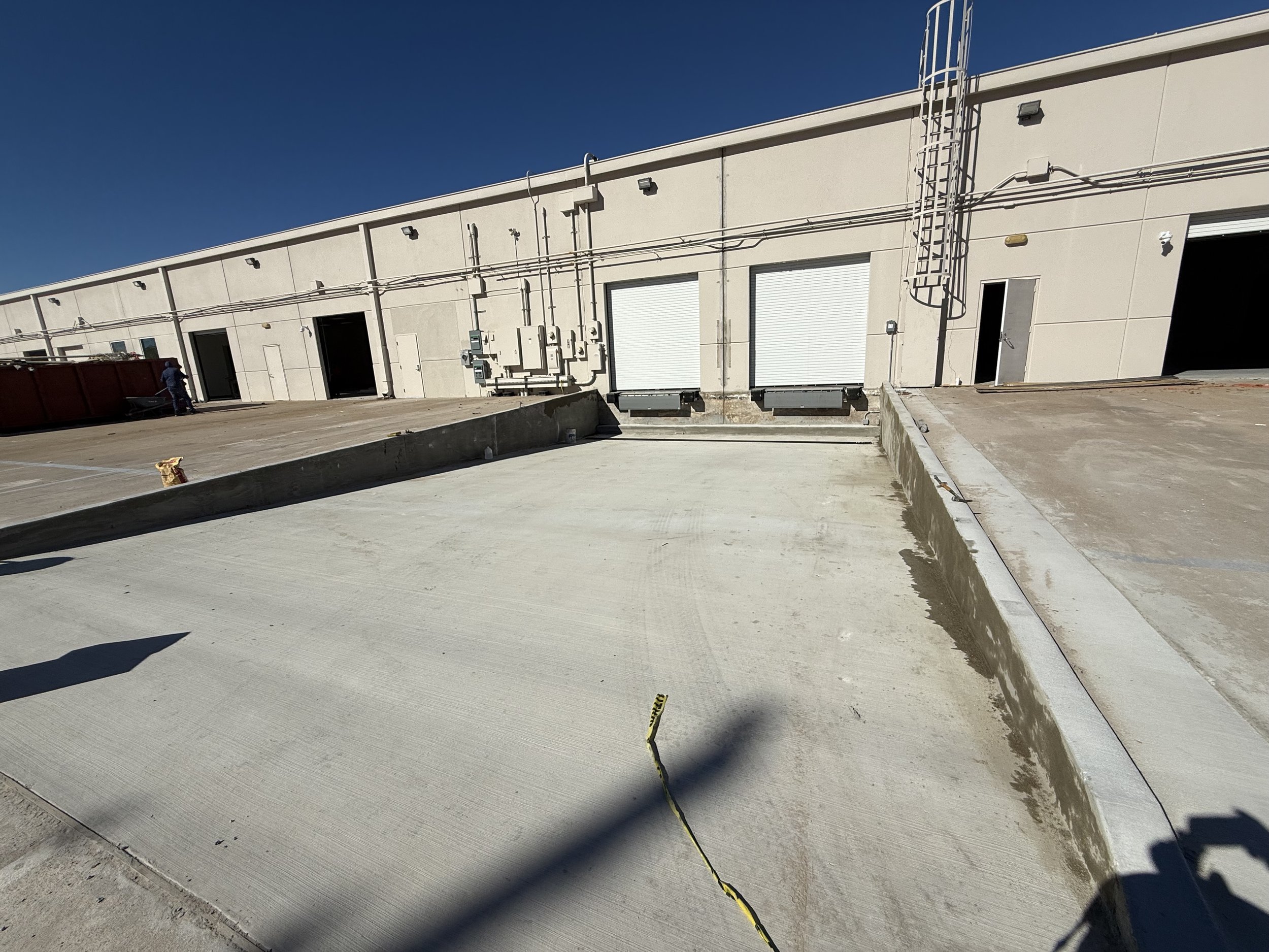 An industrial building with multiple garage doors, electrical meters, and pipes on the exterior wall. A concrete driveway leads up to the building, with some construction materials and a person visible in the distance.