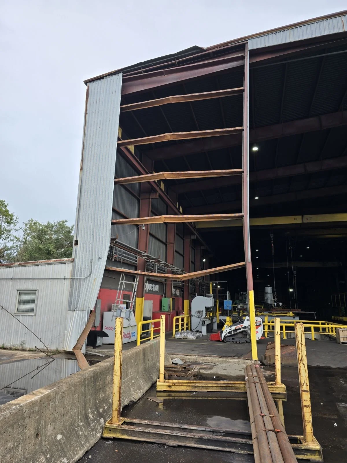 Construction site with a partially built large metal warehouse, exposed wooden beams on the upper levels, and machinery on the ground floor. Yellow safety barriers and pipes are also visible.
