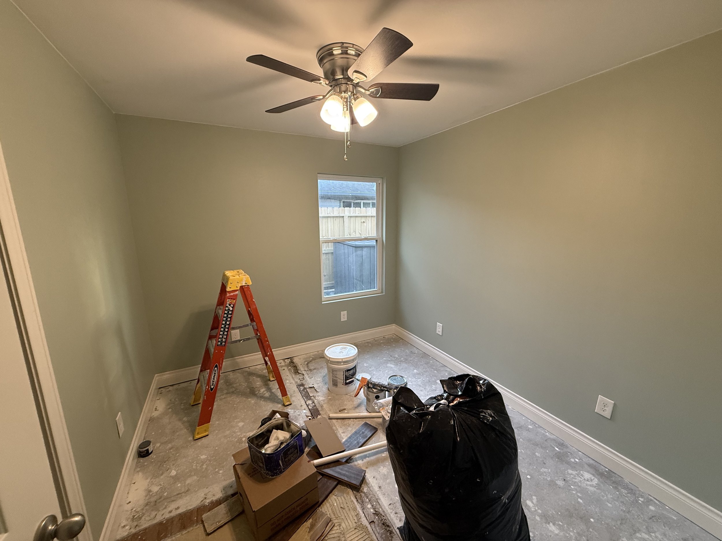 Empty room with pale green walls, window, ceiling fan, and construction supplies including a ladder, buckets, bags, and tools on the floor.