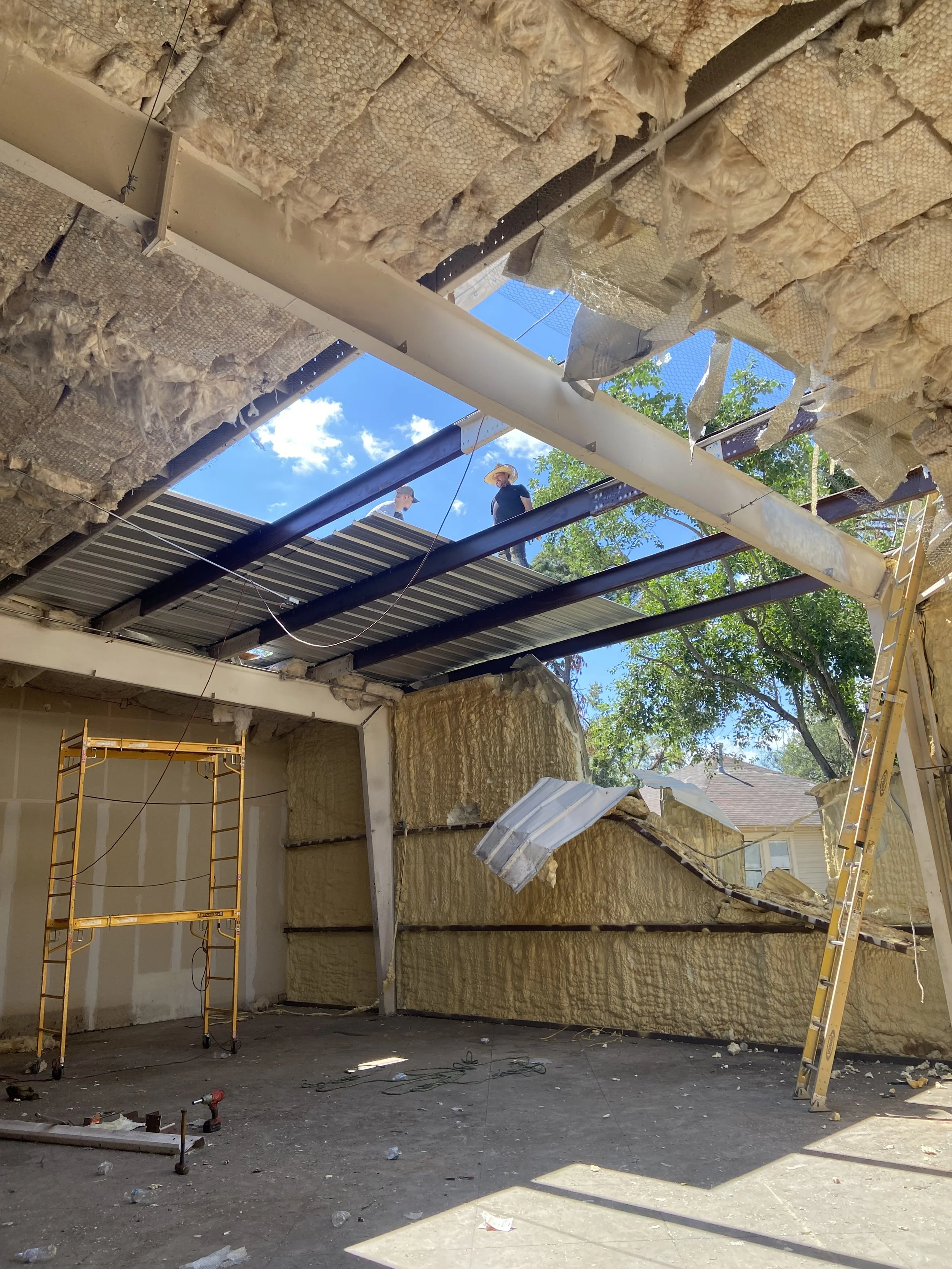 Construction workers are working on a building with an incomplete wall, insulation material on the walls, and a partially built roof with exposed steel beams and a roof panel lifted to reveal the blue sky and trees outside.