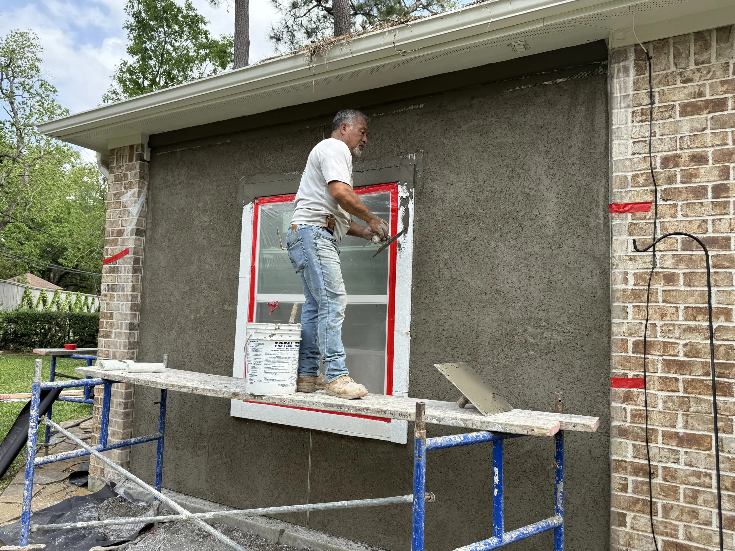 A man standing on scaffolding, applying gray stucco to the exterior wall of a house, with a bucket of stucco nearby.