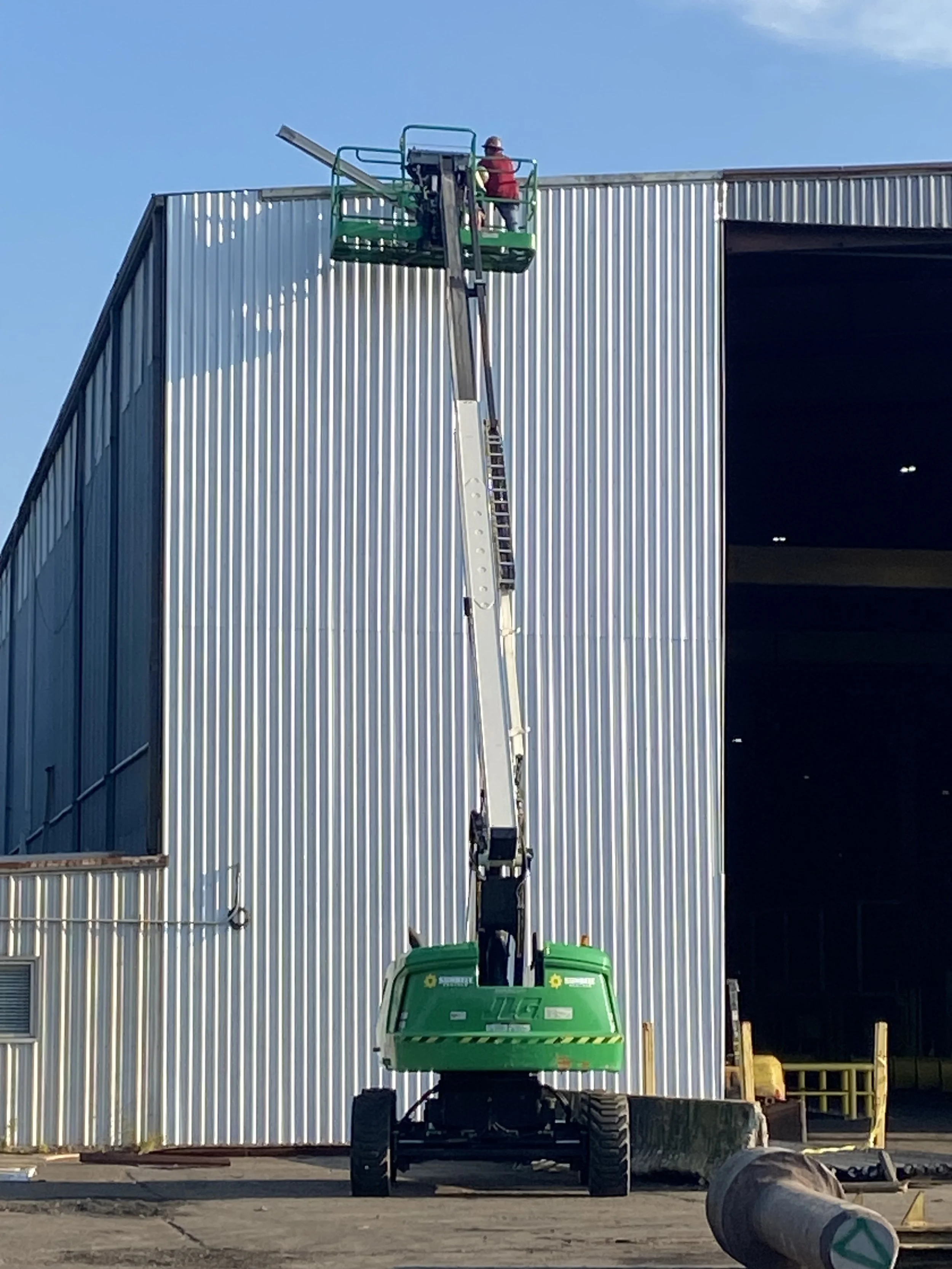 A worker in a red shirt and hard hat operating a green cherry picker lift, working on the upper part of a corrugated metal building under a blue sky.