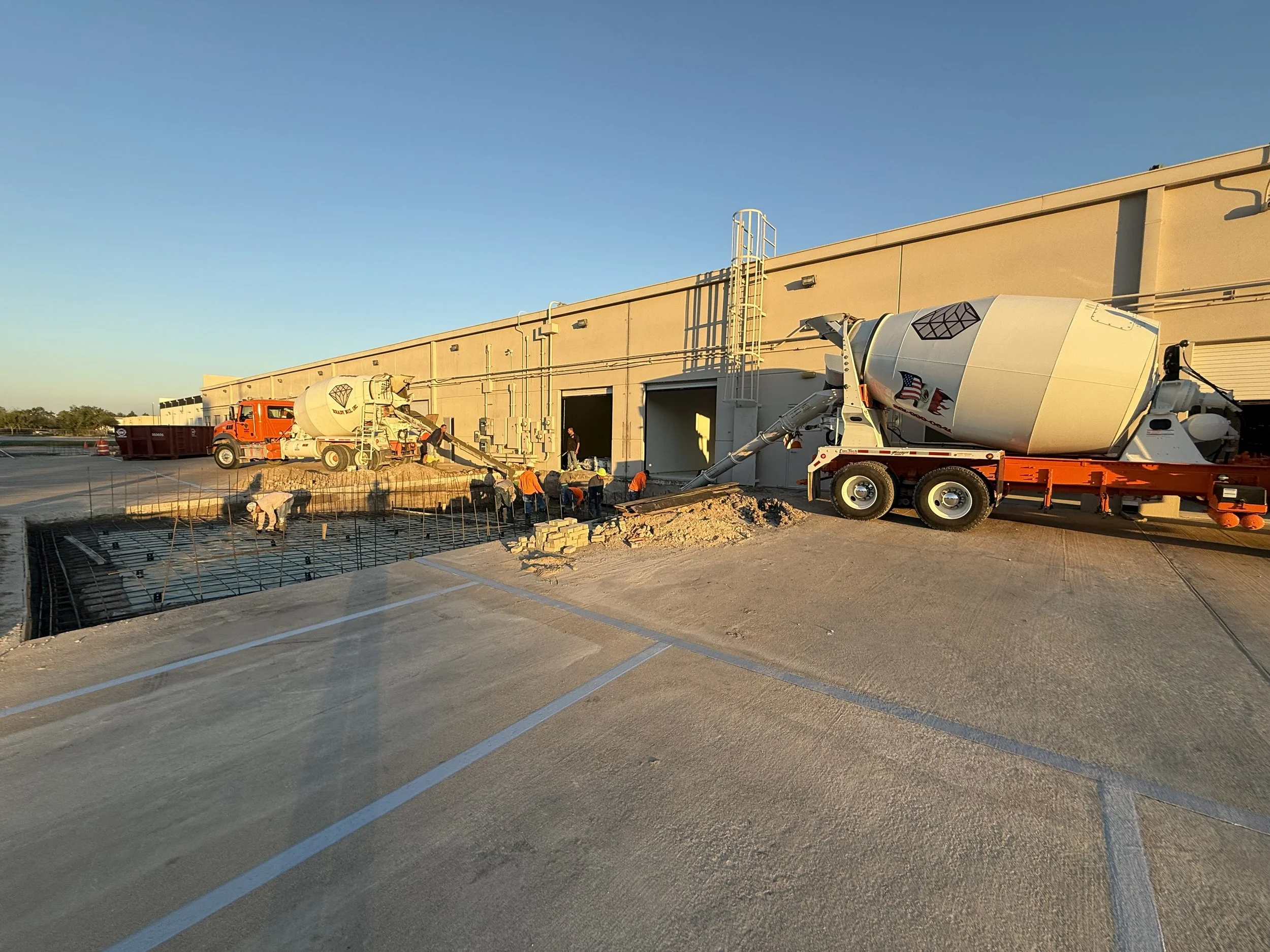 Construction workers pouring concrete using a cement mixer truck outside a large building at sunset.