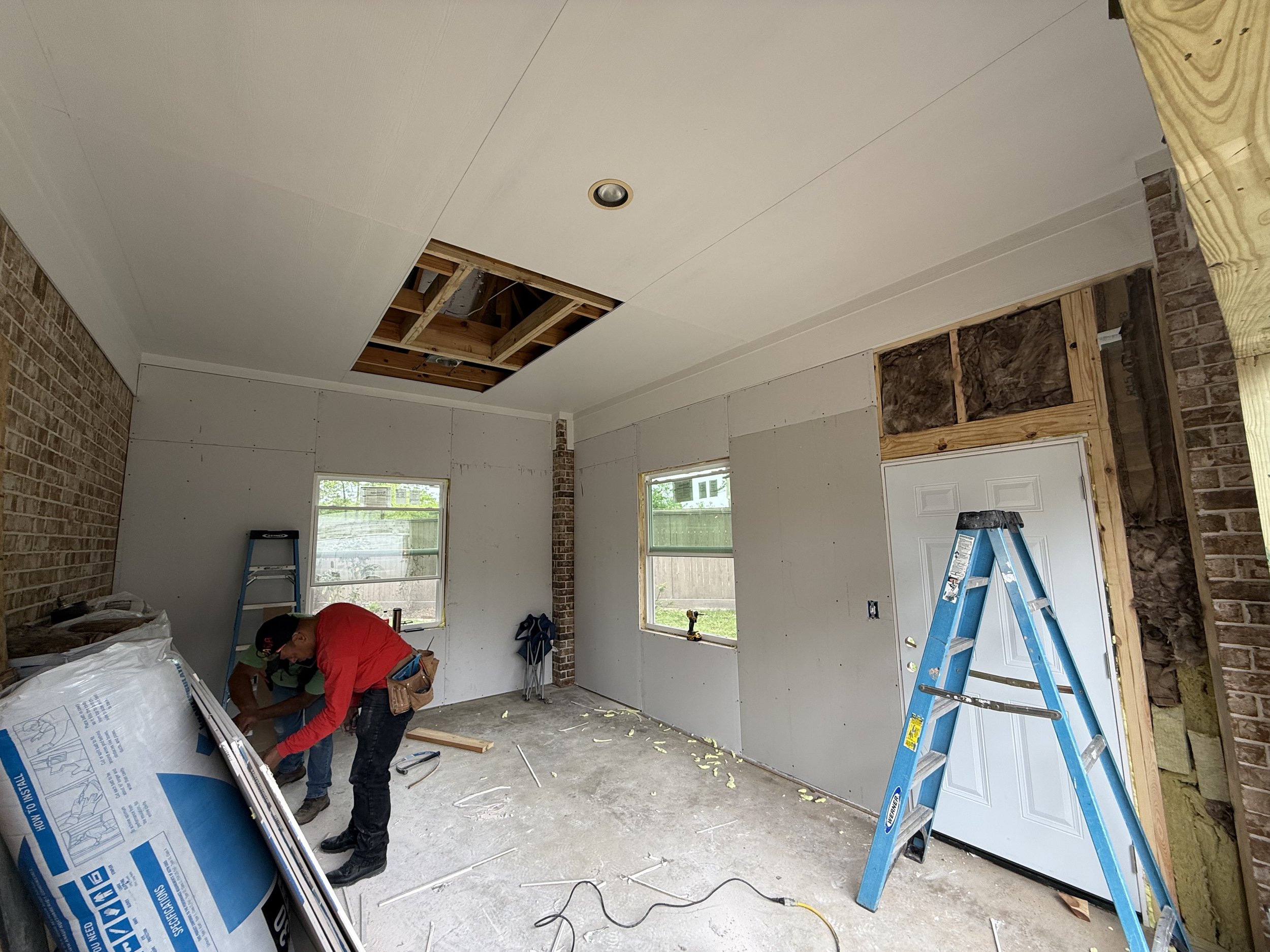 Interior of a room under renovation with drywall installation, a worker in a red shirt working, a ladder, a house door, open ceiling with insulation, and construction materials scattered on the floor.