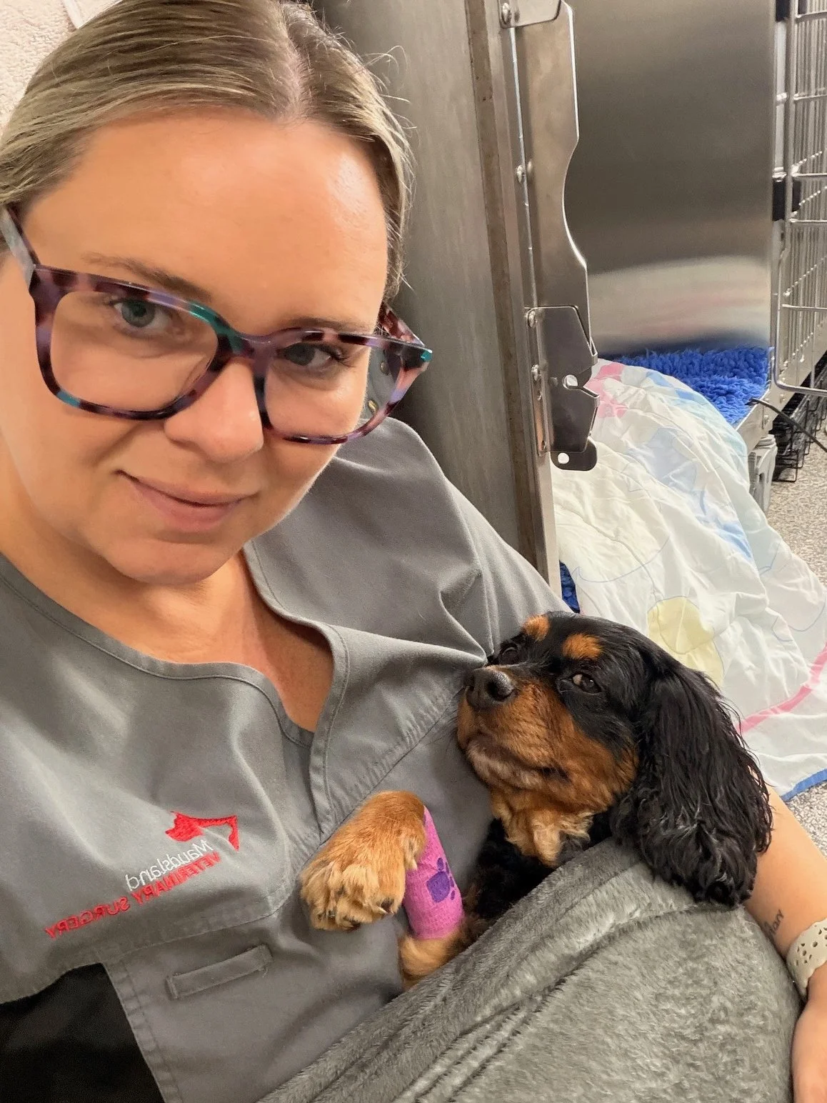 A woman with glasses wearing a vet scrubs holding a small black and tan puppy with a purple bandage on its paw, in a kennel room.