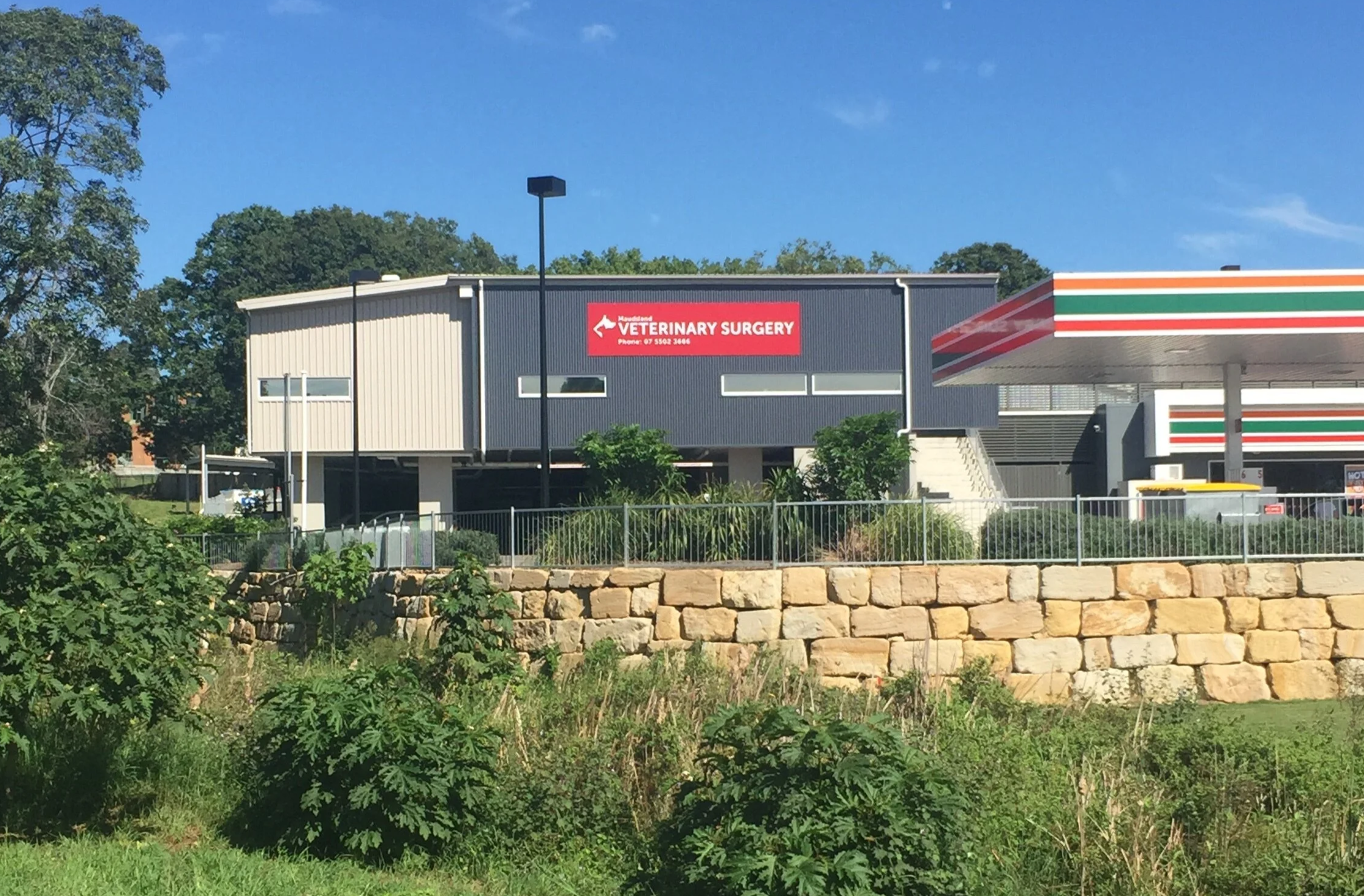 A commercial building with a sign for veterinary surgery, next to a 7-Eleven convenience store, with greenery and a stone wall in the foreground, under a clear blue sky.