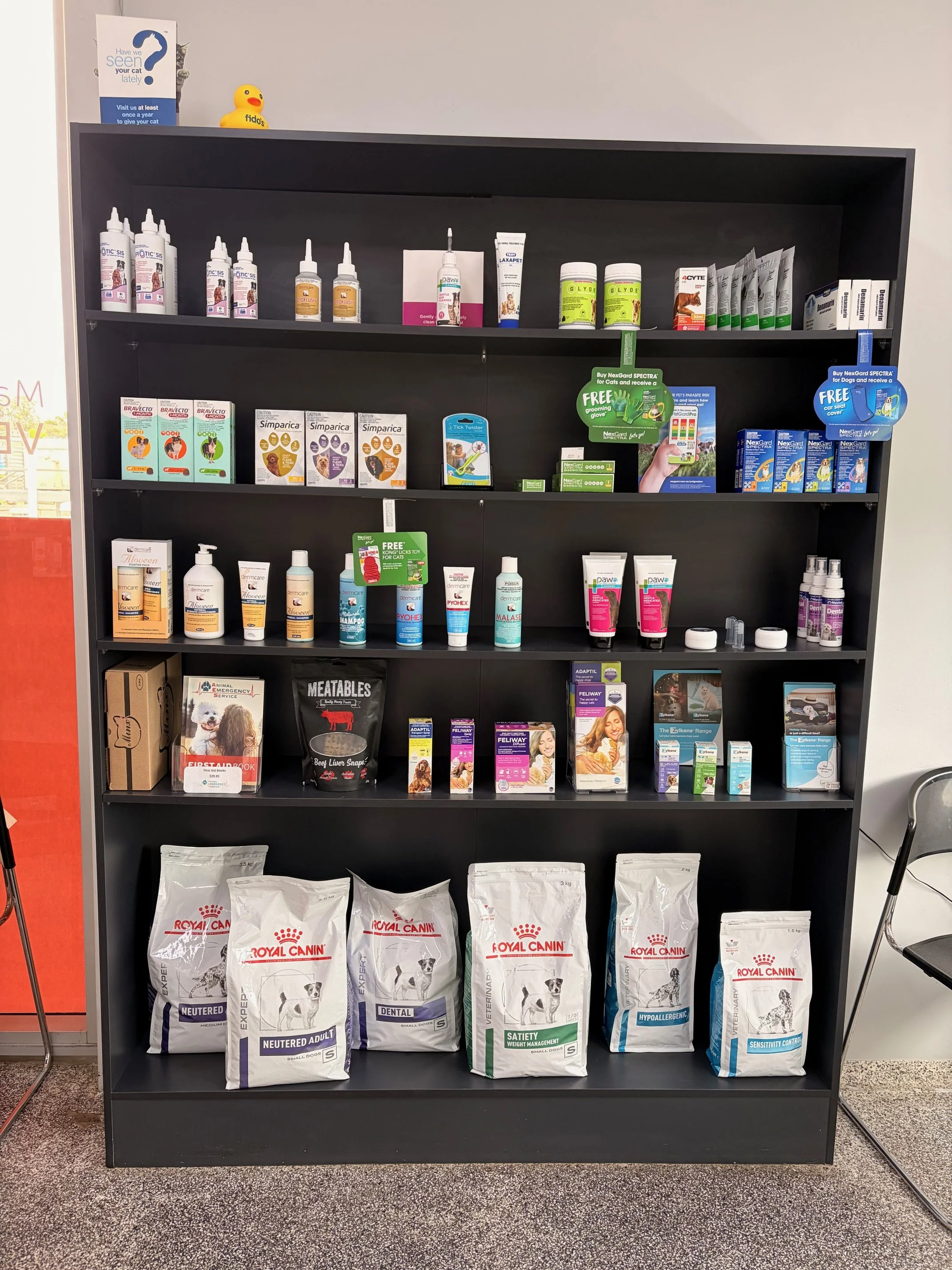 Overlay of pet care products on a black shelf, including pet foods at the bottom, grooming supplies, medications, and accessories at the top, with a yellow rubber duck on top and a fitness poster on the wall.