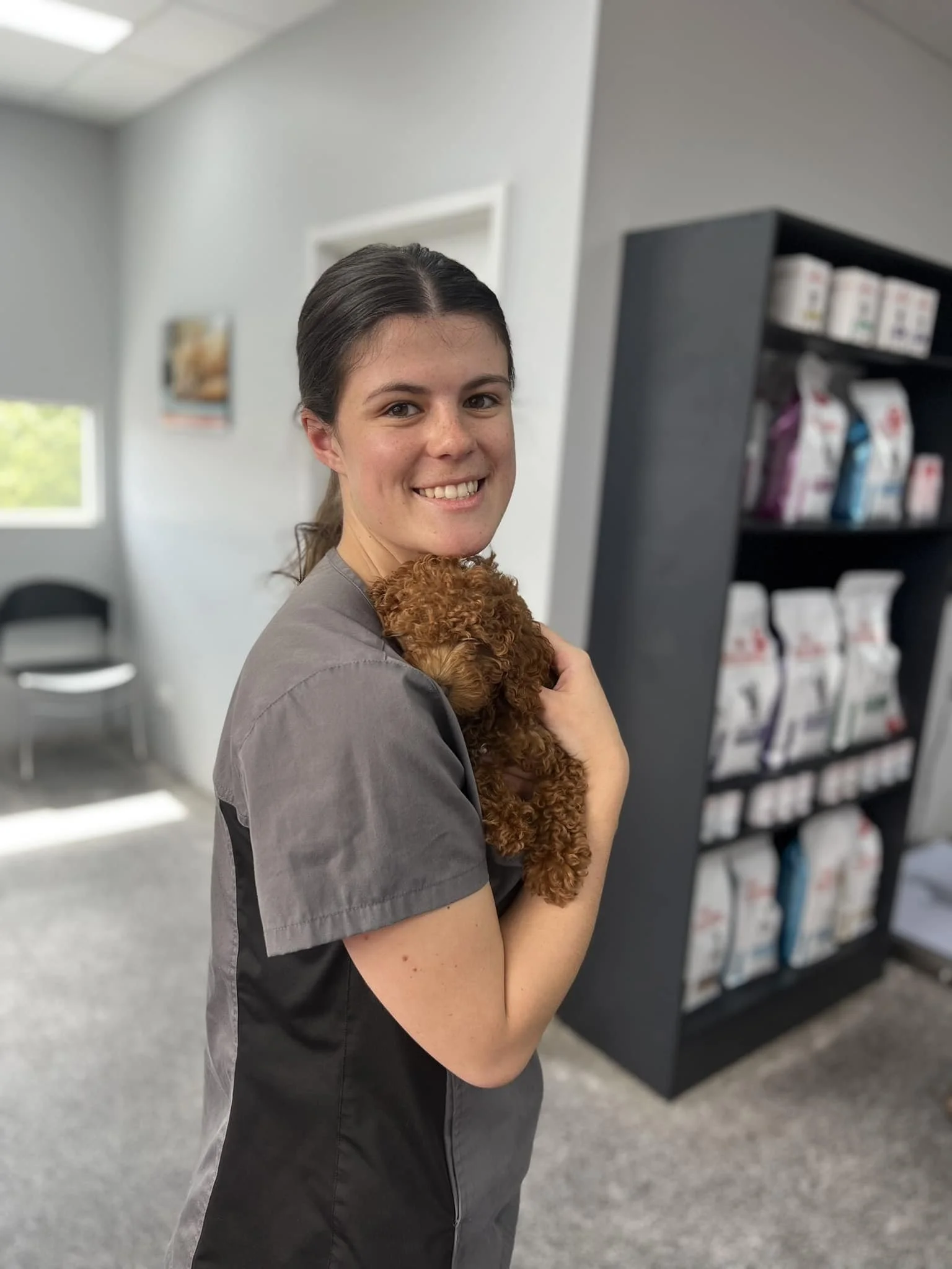 A young woman with dark hair smiling and holding a small curly-haired brown puppy in an indoor veterinary clinic or pet store.