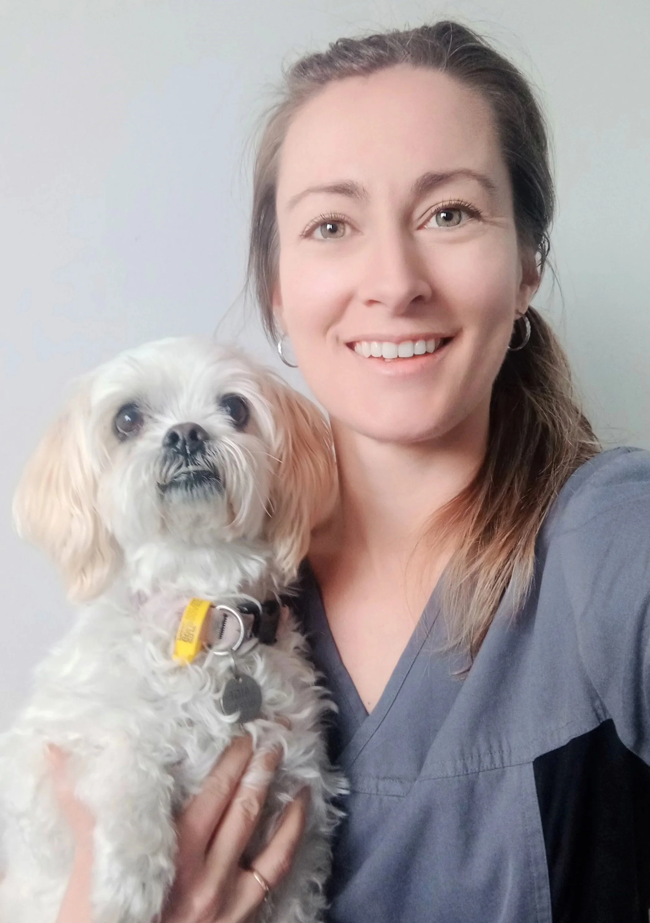 A woman in medical scrubs smiling while holding a small white dog with tan ears and black eyes.