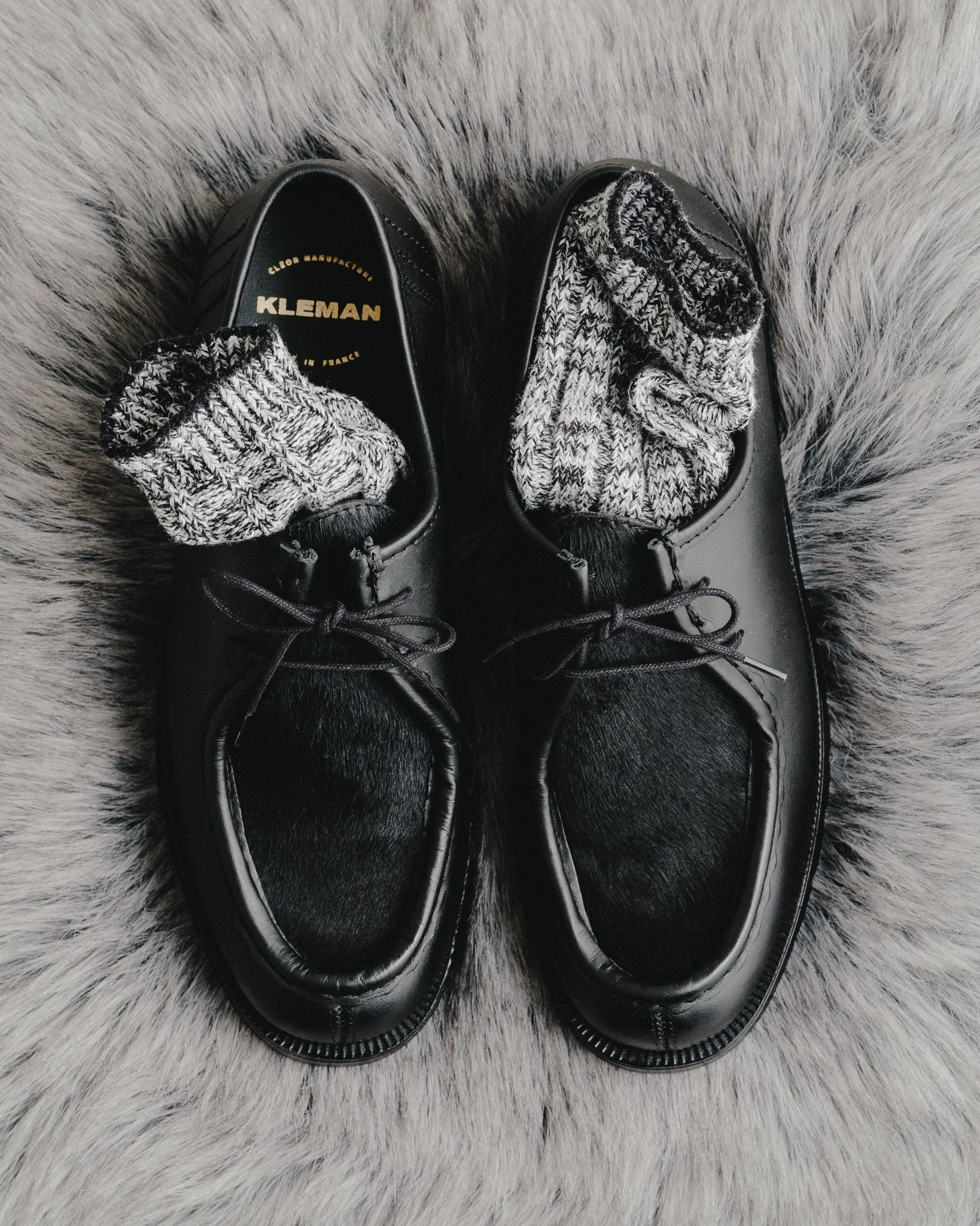A pair of black leather shoes with gray and white knit socks, placed on a gray furry surface.