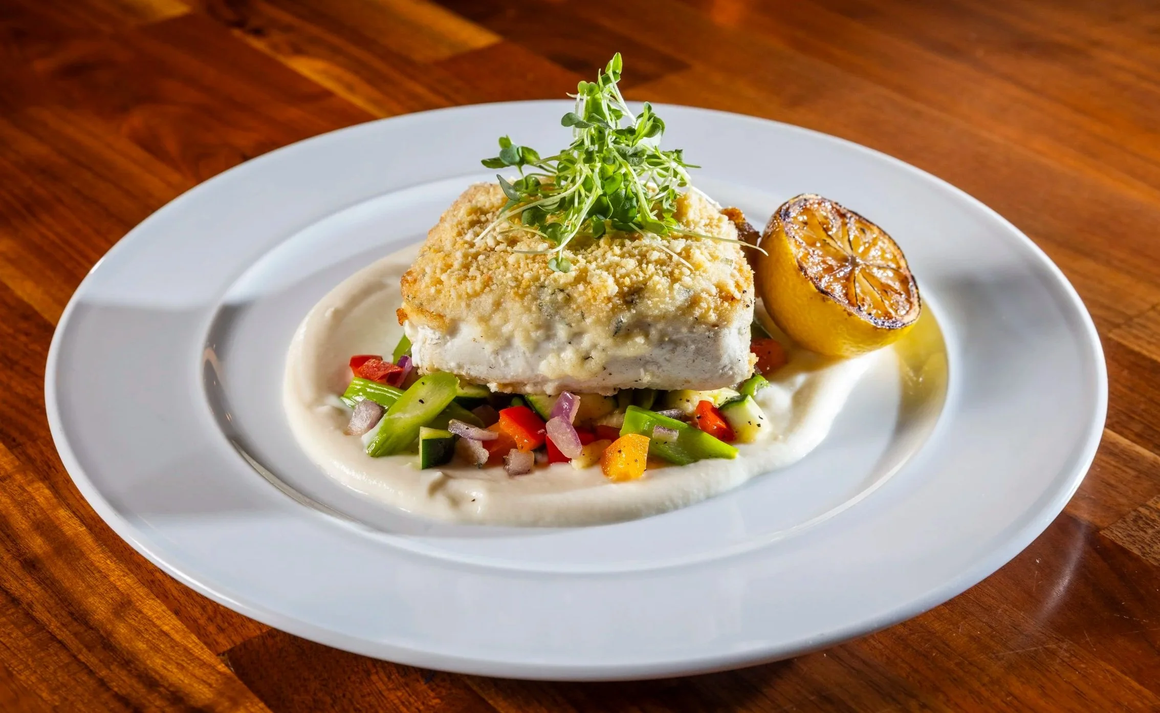 A plated dish featuring a piece of parmesan-crusted halibut and microgreens, accompanied by Seasonal vegetable sauté, cauliflower purée, and charred lemon on a white plate set on a wooden table.