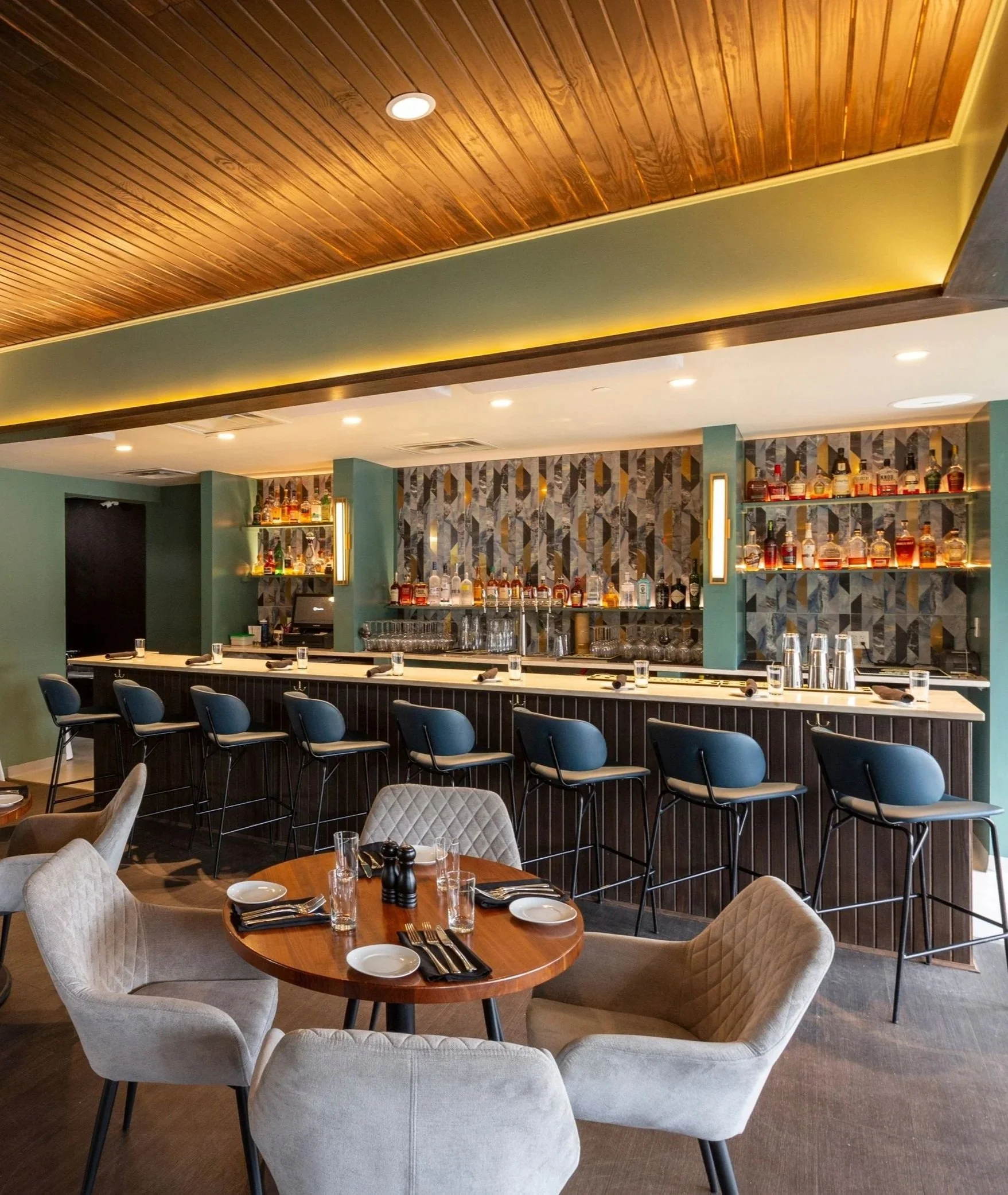 A bar with a white marble counter and a display of liquor bottles behind it, with eight blue barstools in front. There is a round table with four gray upholstered chairs and set tableware in the foreground.