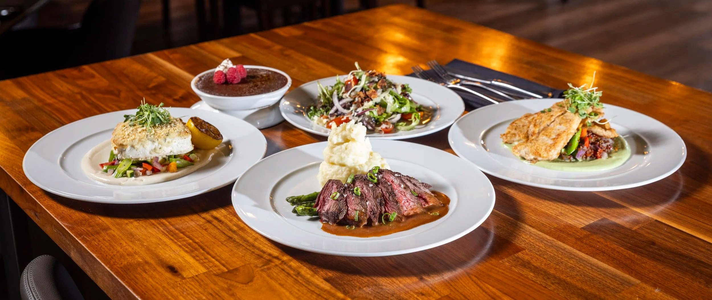 Assorted plated dishes and a dessert on a wooden table in a restaurant setting.