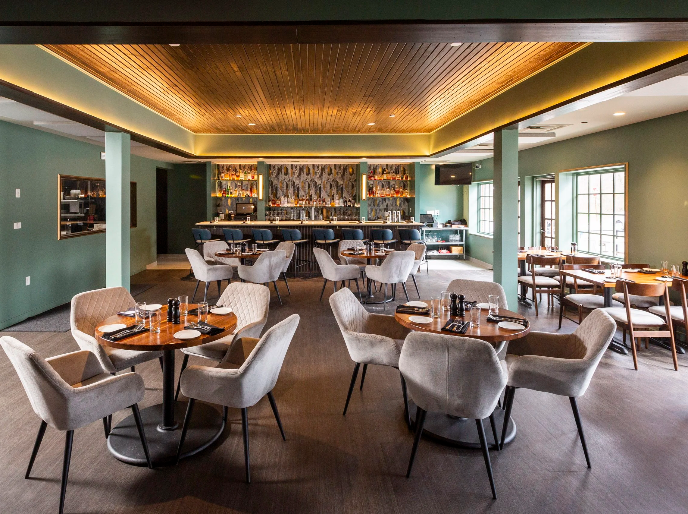 Elegant restaurant interior with round tables, comfortable chairs, and a well-stocked bar area with bottles behind the counter, large windows allowing natural light, and green walls with wood accents.