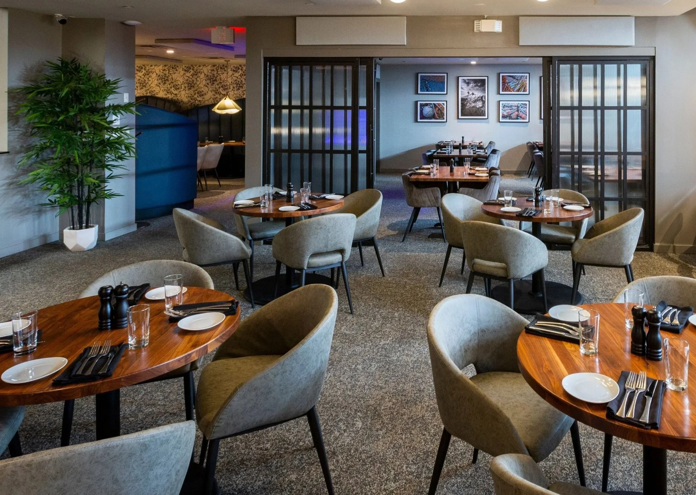 Interior of a modern restaurant with round wooden tables set with plates, glasses, and black napkins, surrounded by beige upholstered chairs, with a large potted plant on the left and framed artwork on the back wall.