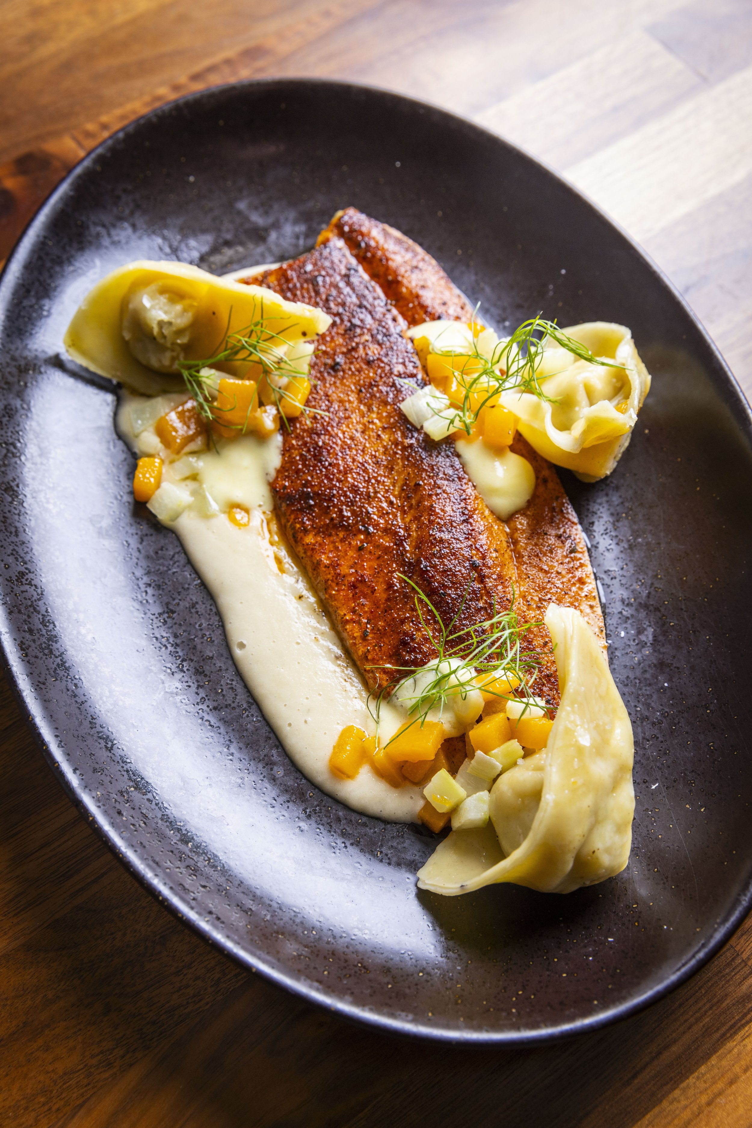 A plated dish featuring blackened rainbow trout, soubise, fresh lemon curd, sweet potato, fennel, and smoked trout ravioli on a dark, oval plate.