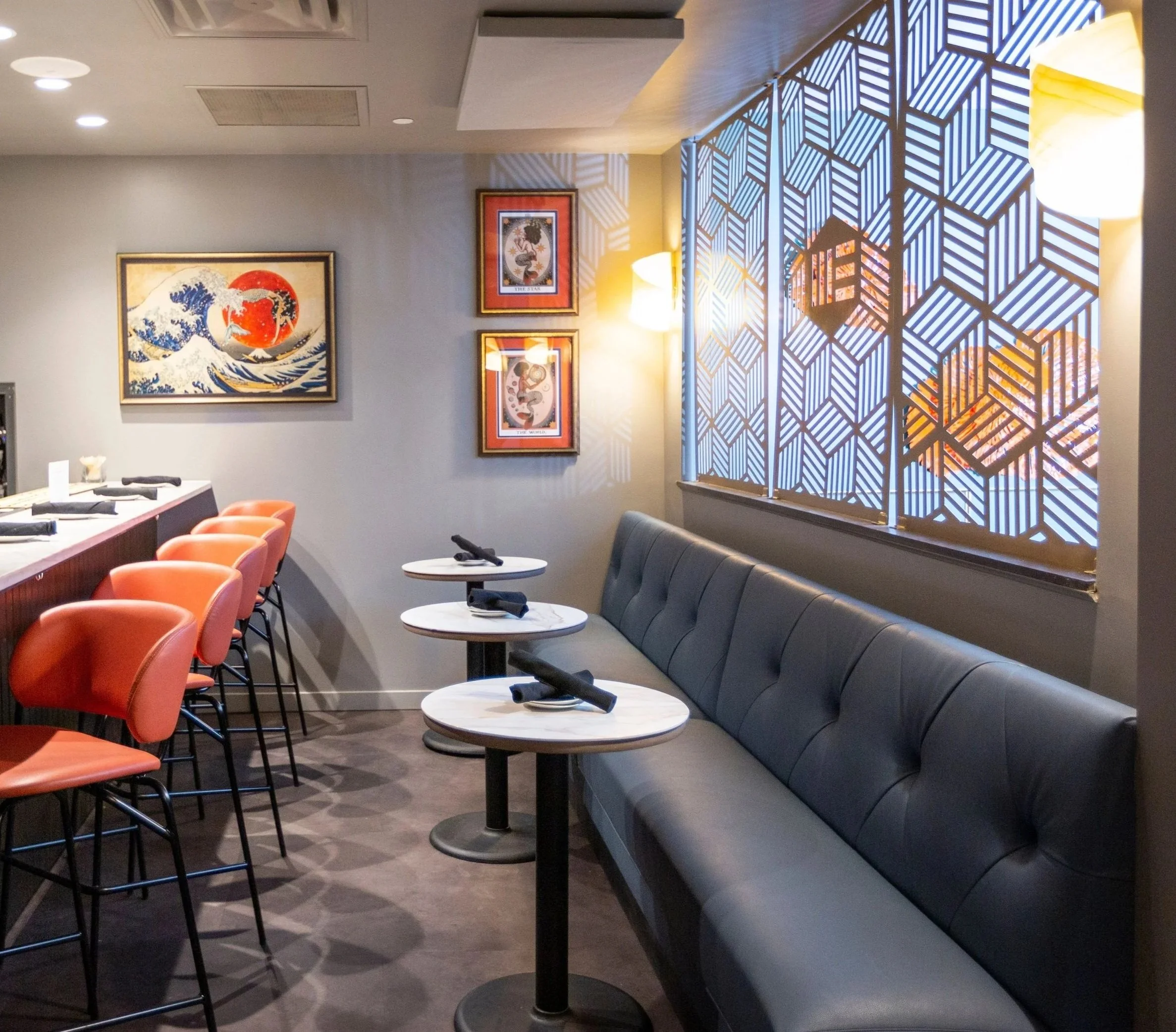 A cozy restaurant corner with a black leather banquette, three small round tables set with black napkins, a colorful painting of a wave, and two framed posters on the wall. A decorative window with geometric patterns casts shadows, and soft yellow wall lights illuminate the space.