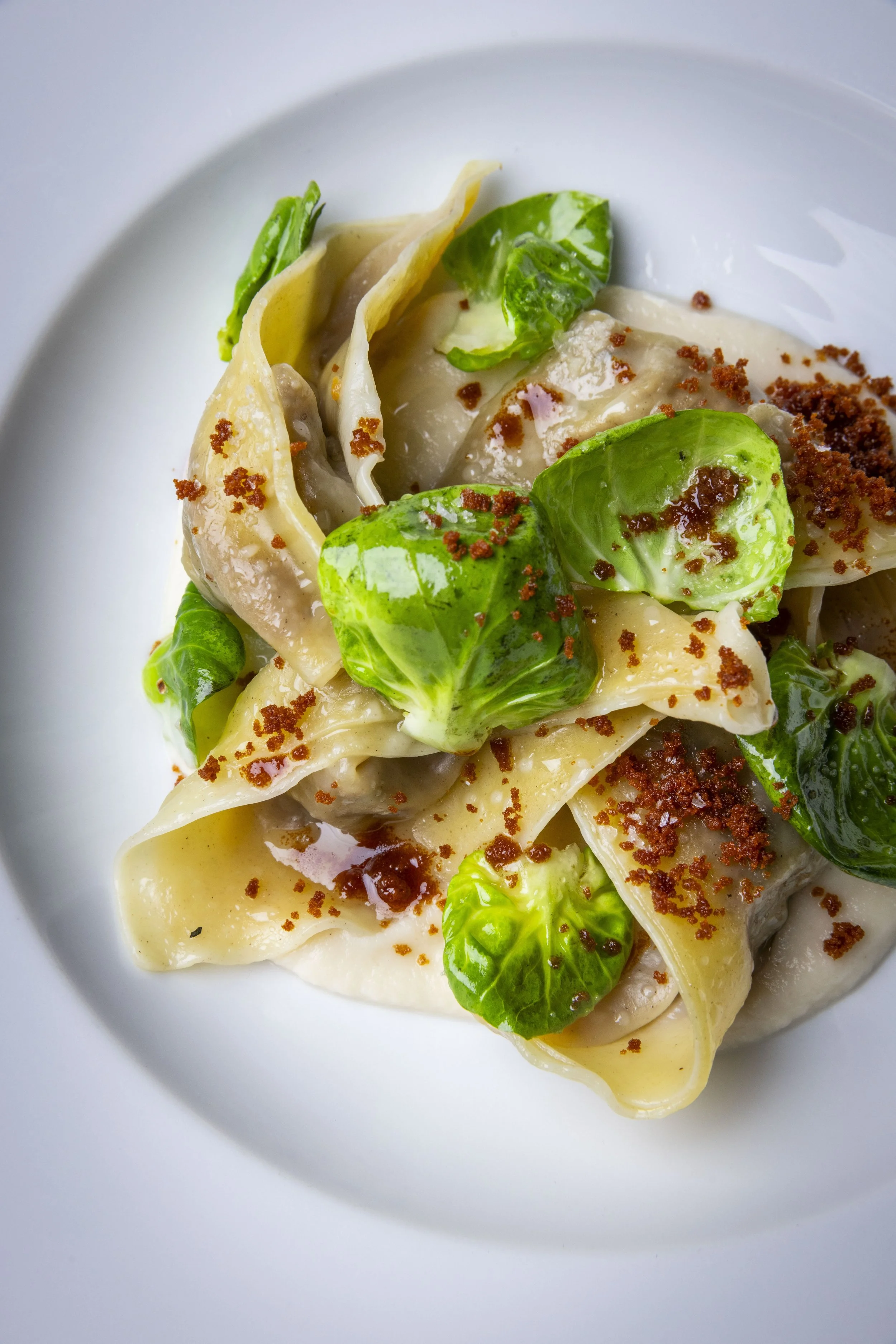 Close-up of a plate of ravioli pasta with cauliflower purée, Brussels sprout leaves, brown butter crumble, microgreens, and truffle oil.