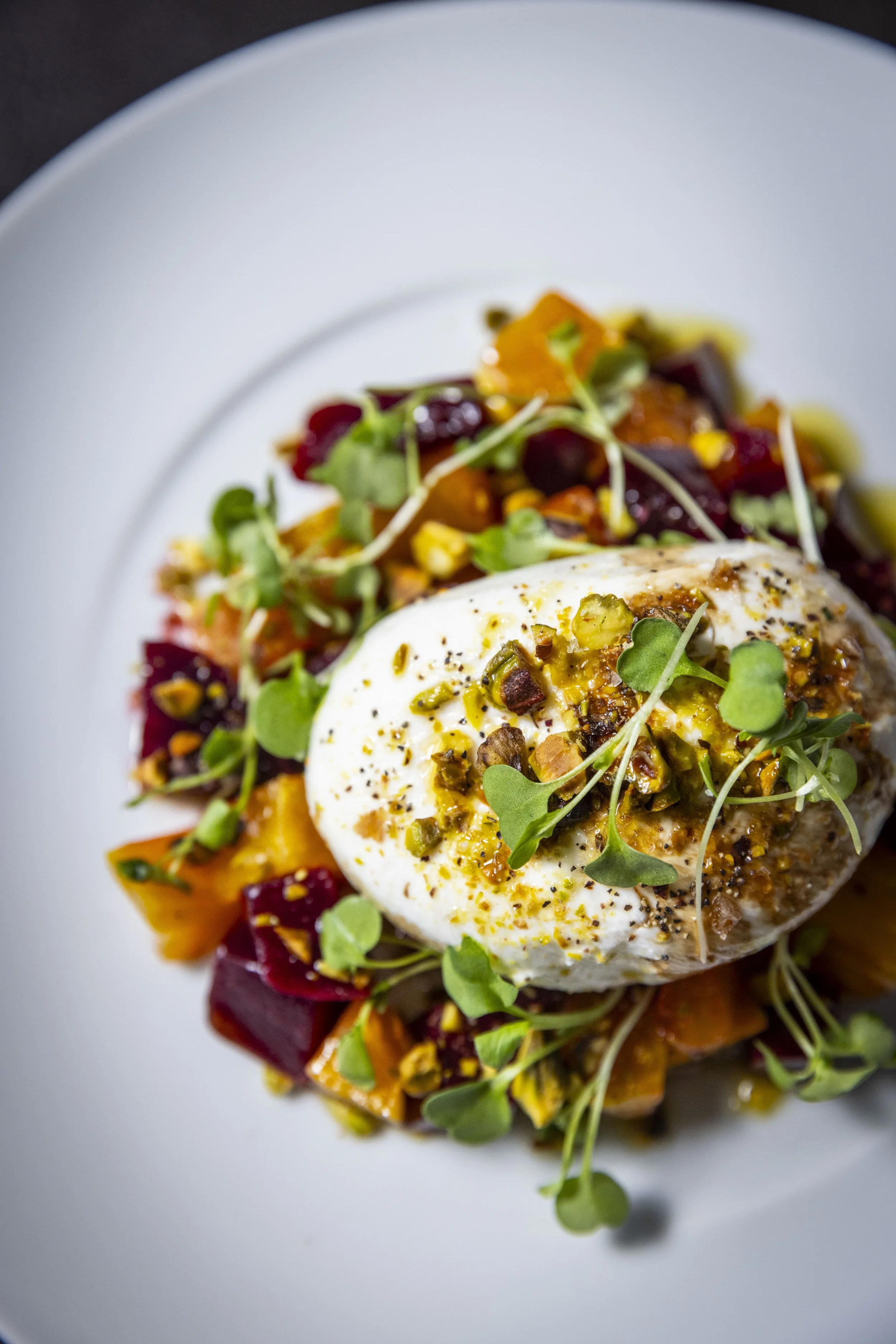 A dish featuring burrata topped with red & golden beets, horseradish-sherry vinaigrette, pistachios, arugula, balsamic, and Extra Virgin Olive Oil on a white plate.