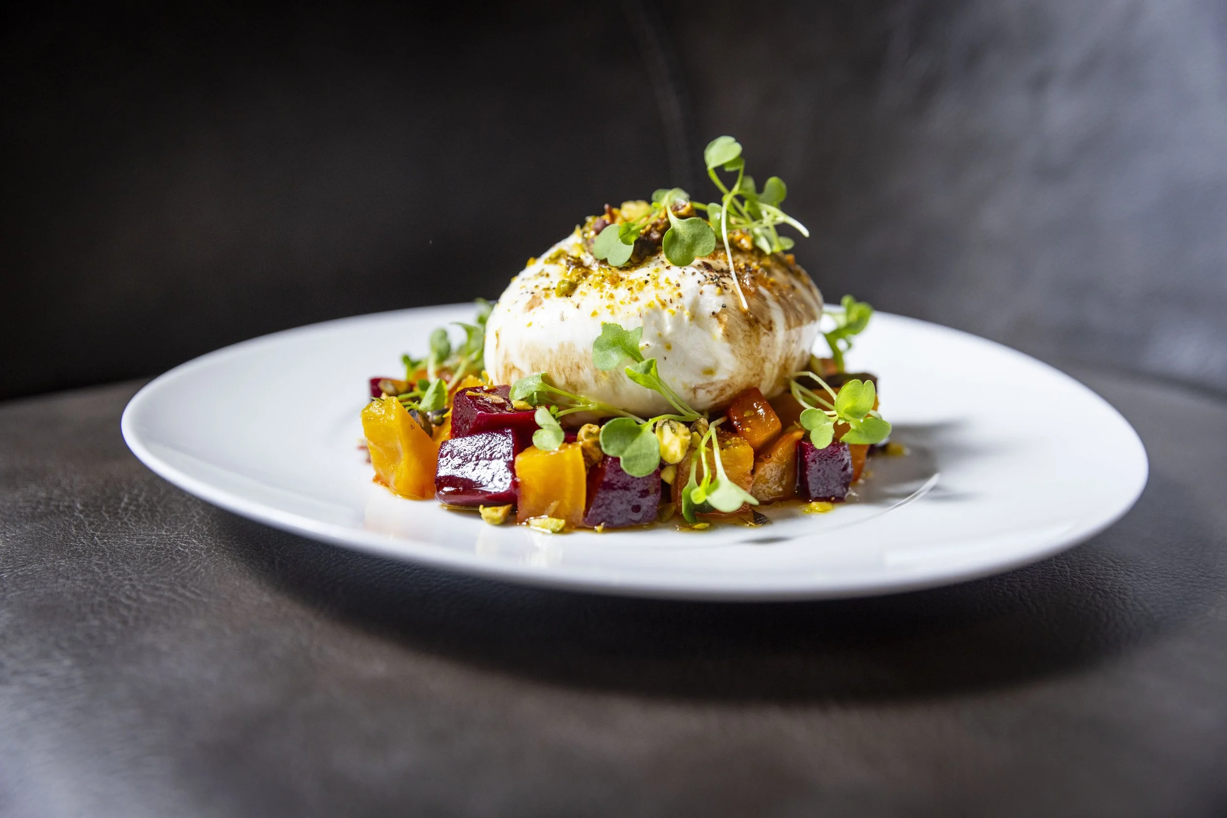 A gourmet dish featuring a burrata topped with microgreens, served on a bed of red and golden beets on a white plate against a dark background.