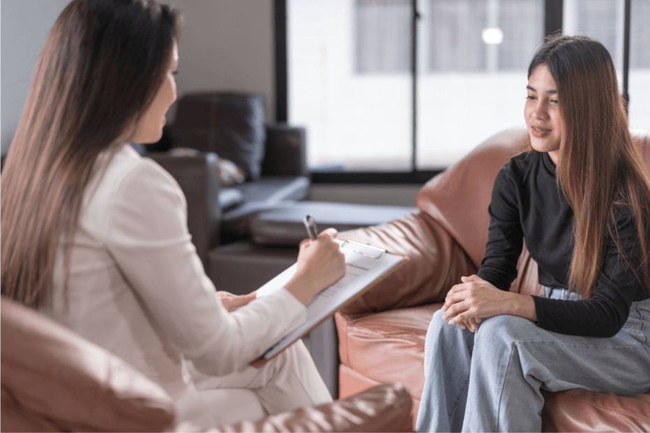 woman-sitting-on-brown-leather-couch-smiling-and-facing-therapist-with-clipboard-in-bright-relaxed-setting-during-counseling-session-paraclete-counseling-center