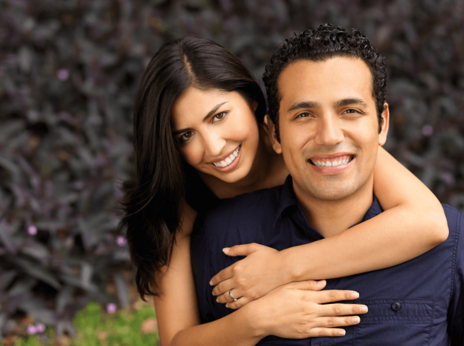A smiling couple stands outdoors as the woman with long dark hair hugs the man in a navy shirt from behind, surrounded by lush greenery.