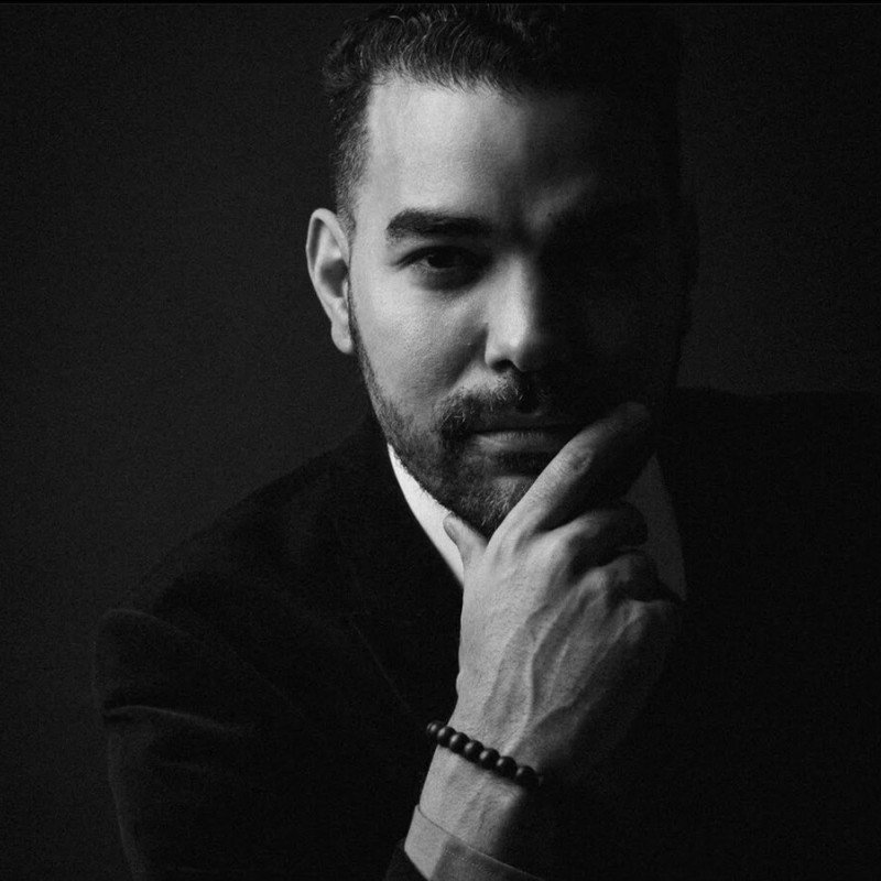 A black and white close-up portrait of a man with a beard, dressed in a suit, with his hand resting on his chin, looking contemplative.