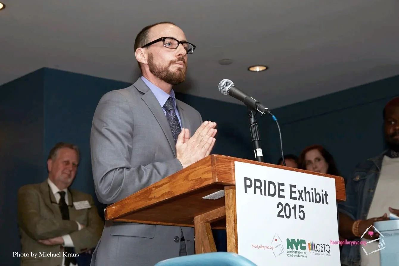 Man in gray suit and glasses speaking at a podium during a Pride Exhibit event in 2015, with several people in the background.