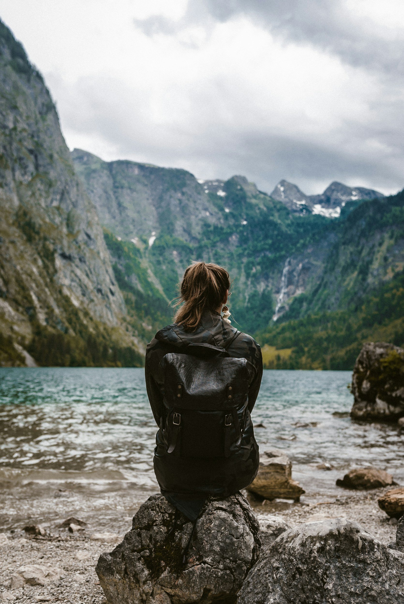 A person with brown hair, wearing a black jacket and backpack, sitting on a rock by a lake with mountains and cloudy sky in the background.
