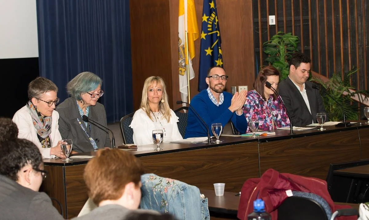Panel of six people seated at a long table, some clapping, in a conference room with flags and plants in the background.