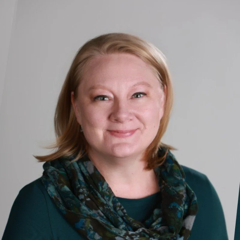portrait of a middle-aged woman with blonde hair wearing a dark teal top and a patterned scarf, smiling at the camera