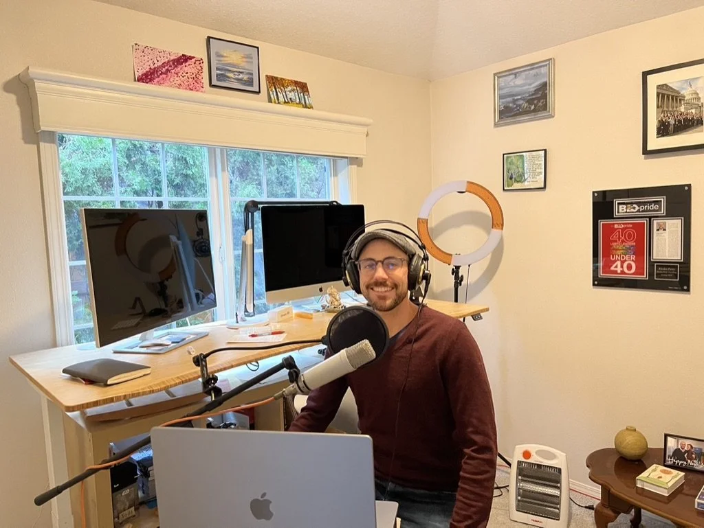 A man sitting at a desk in a home recording studio, smiling at the camera, wearing headphones and glasses, with a microphone in front of him. The room has two computer monitors, artworks on the walls, and a window with trees outside.