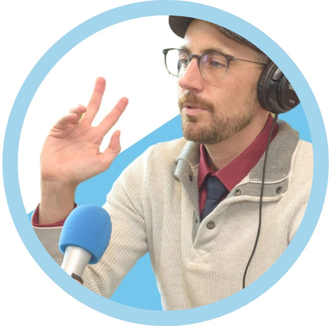 A man wearing glasses, a newsboy cap, and headphones speaking into a blue microphone while gesturing with his hand.