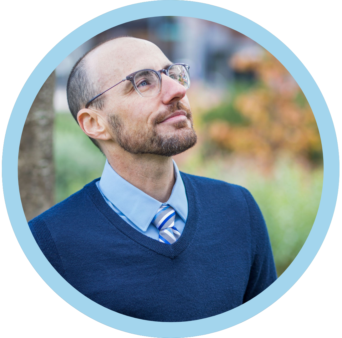 A man wearing glasses, a blue sweater, a light blue dress shirt, and a striped tie, looking upward with a thoughtful expression.