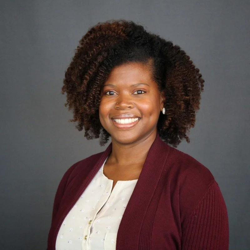 A woman with curly hair smiling against a plain gray background, wearing a white top and maroon cardigan with earrings.