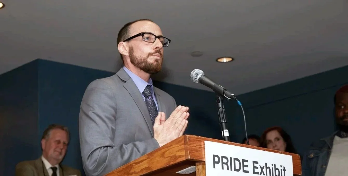 A man wearing glasses, a gray suit, and a tie is standing at a podium with a sign that reads "PRIDE Exhibit." He has his hands clasped in front of him and appears to be speaking or praying. There are several people in the background, including a man in a suit and a woman with dark hair.