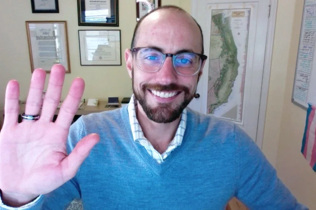 A smiling man with glasses and a beard, wearing a light blue sweater and a checkered shirt, waving at the camera inside an office with framed certificates, a map, and posters on the wall.