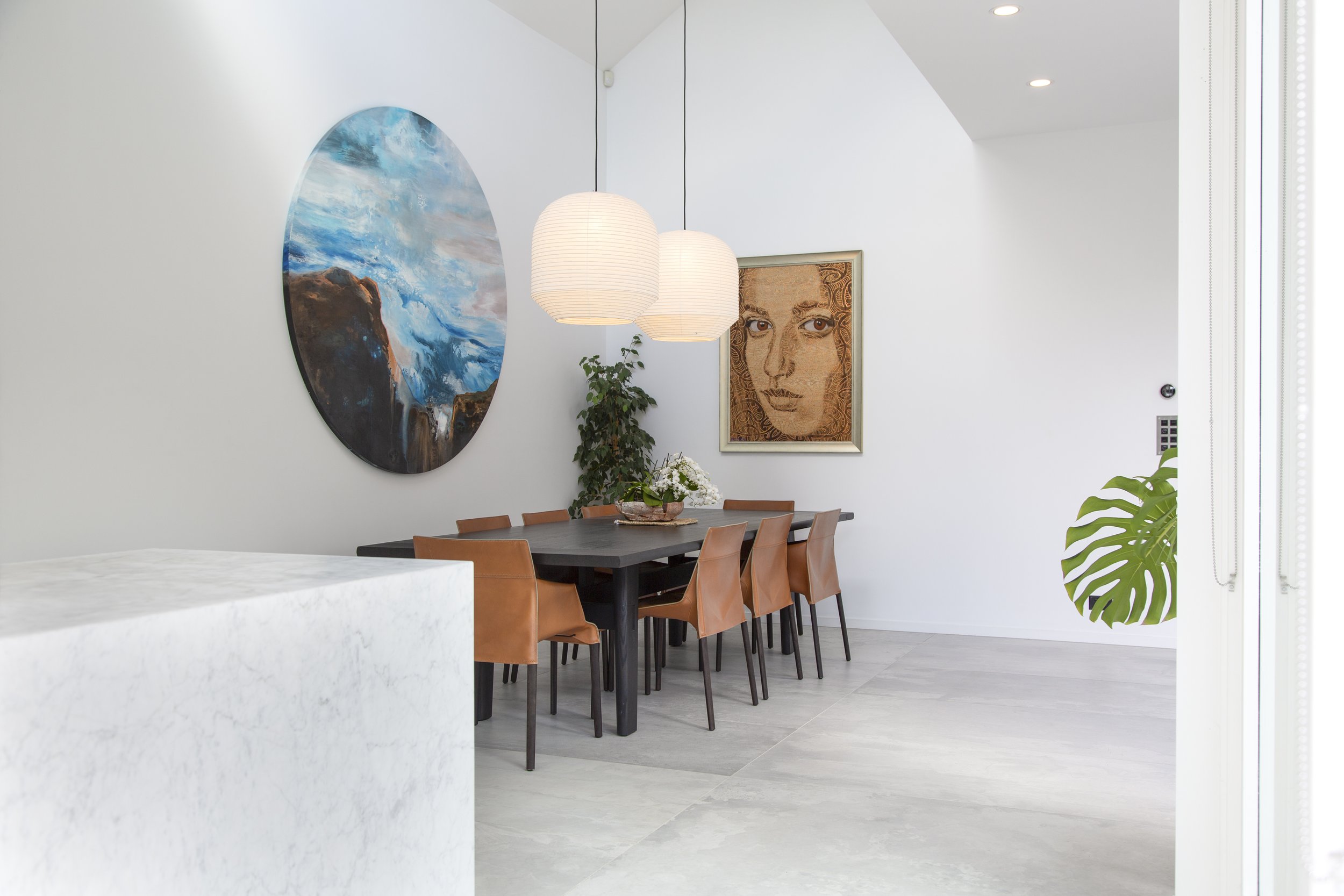Modern dining room with a dark wood table, tan chairs, white walls, and artwork including a large circular painting of ocean waves and a framed portrait of a woman's face. Hanging pendant lights and green plants are also visible.