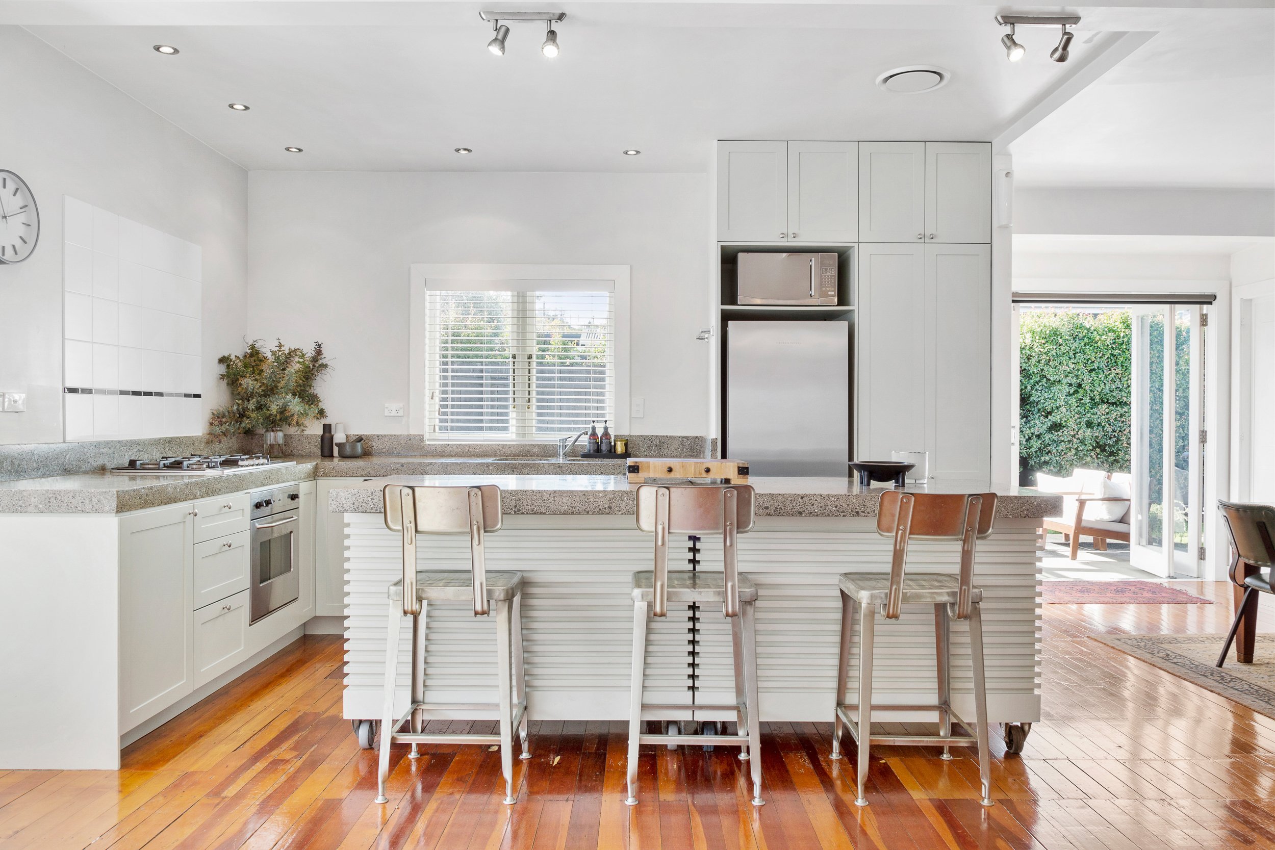 Modern kitchen with white cabinets, stainless steel appliances, a kitchen island with bar stools, hardwood floors, and a sliding glass door leading to the backyard.