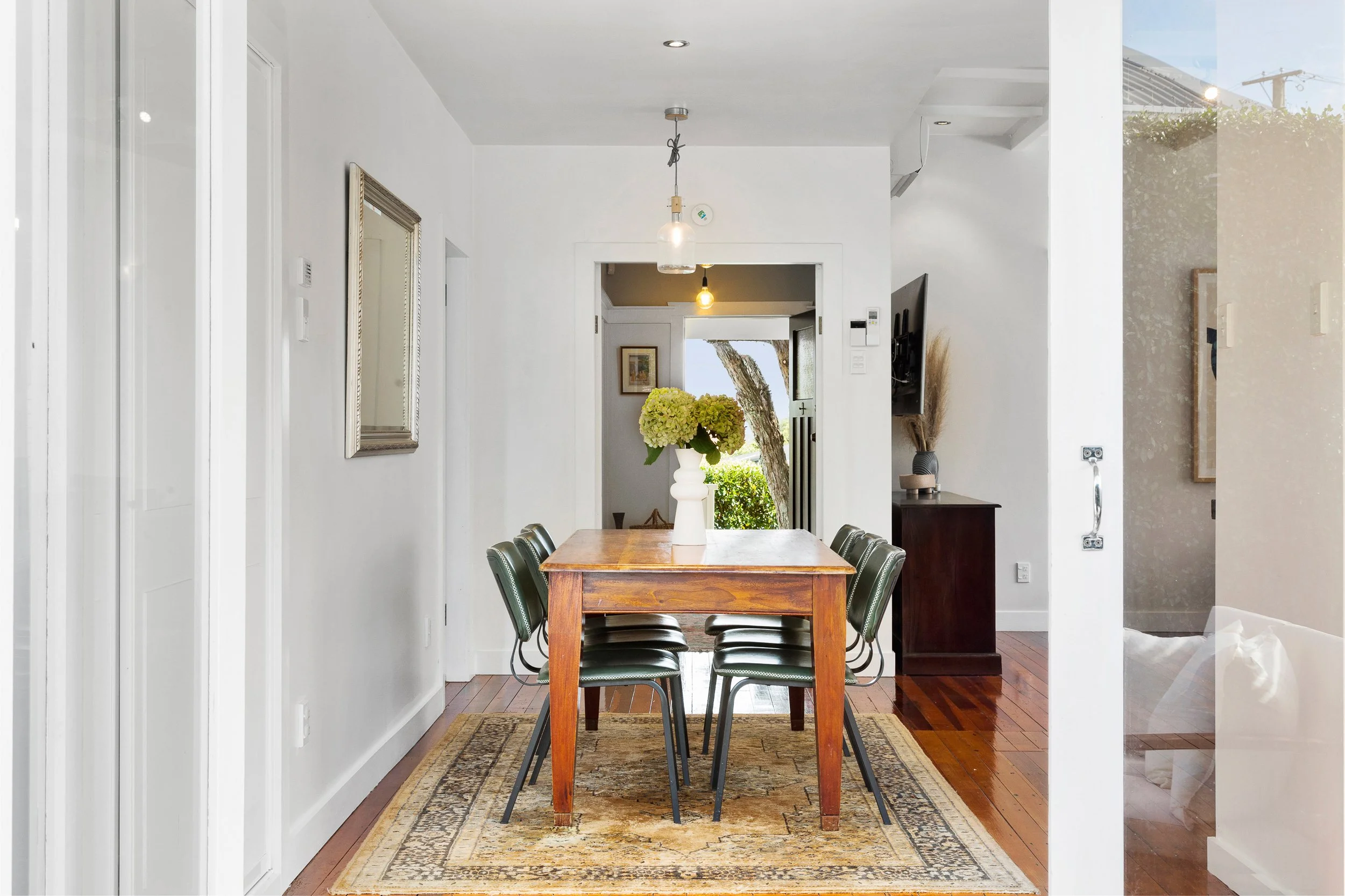 A dining area with a wooden table, black chairs, a vase with green hydrangeas, framed art on the wall, and windows showing trees outside.