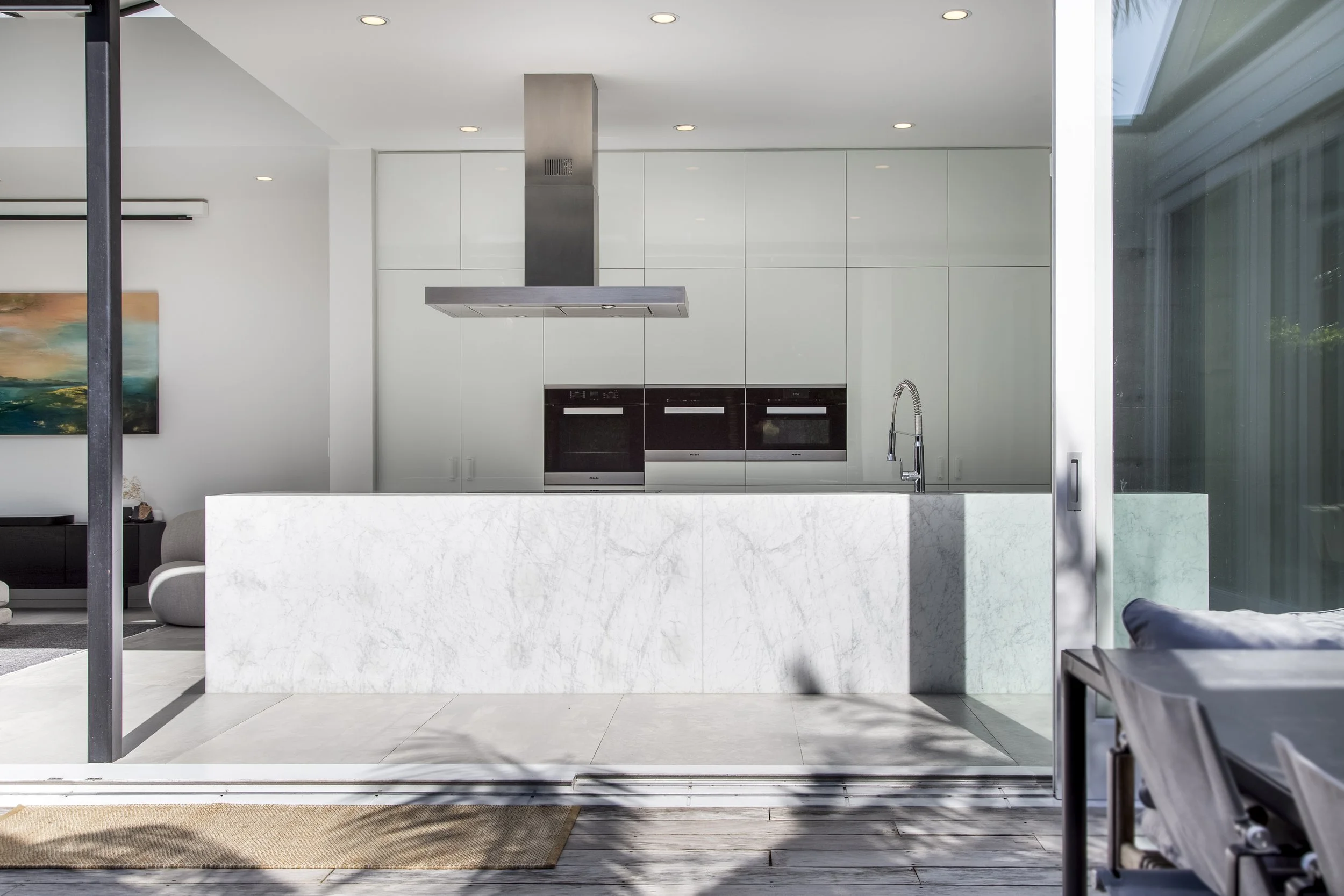 Modern minimalist kitchen with white cabinets, black appliances, stainless steel vent hood, marble kitchen island, and sliding glass door leading outside with patio furniture.