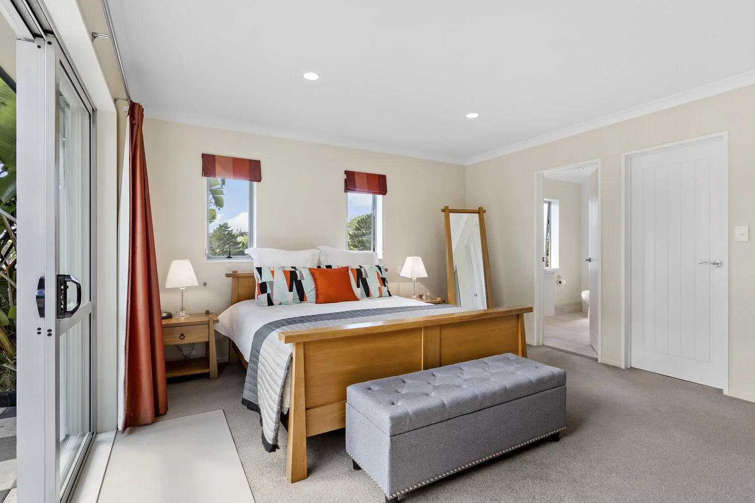 Bedroom with a wooden bed, gray tufted bench, two nightstands with lamps, two windows with red valances, cream walls, and a bathroom visible through an open door.