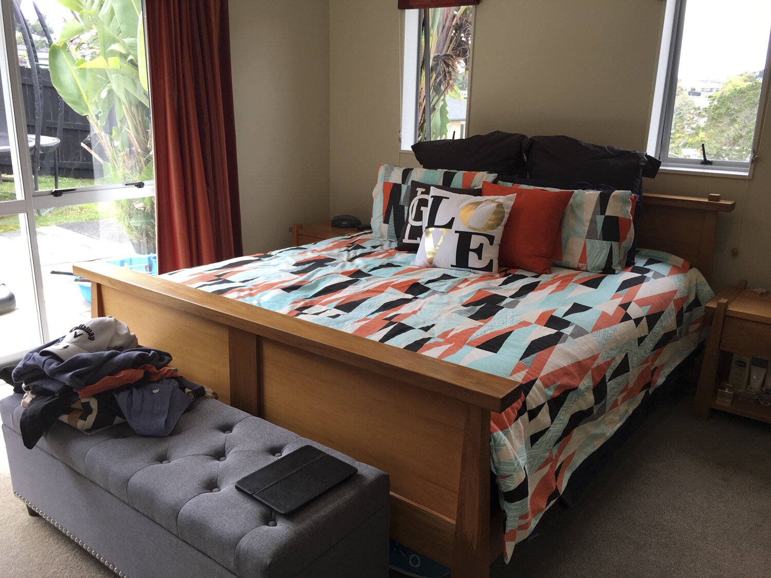 A neatly made bed with a colorful geometric patterned bedspread and decorative pillows spelling "LOVE" in a bedroom. The bed has a wooden frame. There is a sliding glass door on the left with a view of a backyard with plants and outdoor furniture. To the right is a window, and beside the bed is a wooden nightstand with personal items. A gray tufted bench with clothes and a tablet is at the foot of the bed.