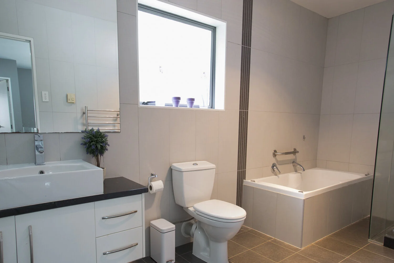 Modern bathroom with white tiled walls, a bathtub with a faucet, a toilet, a sink with a black countertop, a mirror, a window with purple flowers on the sill, and a small potted plant near the sink.