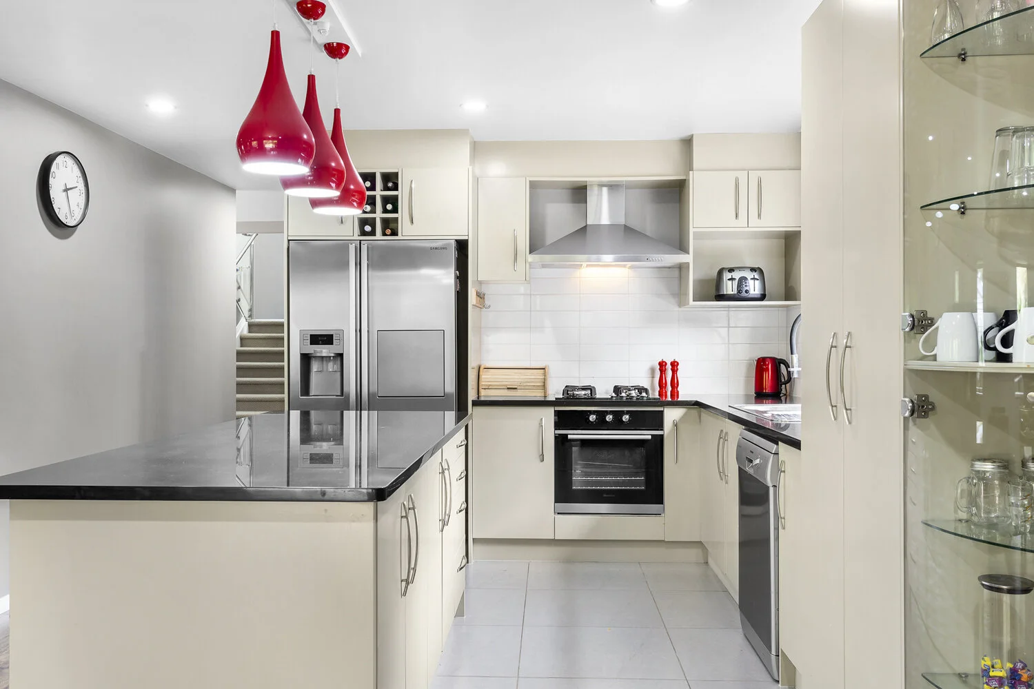 Modern kitchen with beige cabinets, black countertops, stainless steel appliances including a refrigerator, oven, dishwasher, and toaster, with red accents like pendant lights, kettle, and salt and pepper shakers.