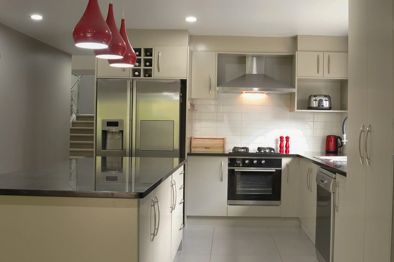 Modern kitchen with stainless steel refrigerator, oven, and appliances, white cabinets, cream tiled backsplash, red accents including hanging pendant lights, kettle, toaster, and salt and pepper shakers.