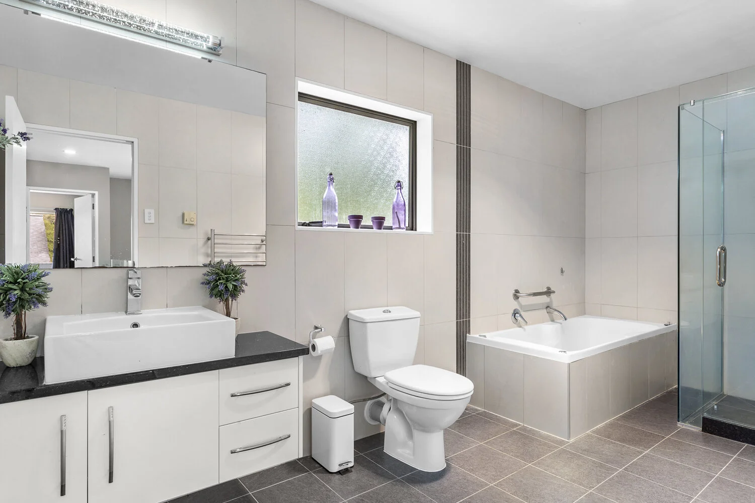 Modern bathroom with a white sink vanity, a mirror, a toilet, a bathtub, and a glass-enclosed shower, with neutral tile and a frosted window.